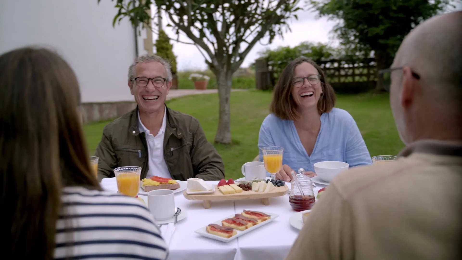 Una familia ríe alrededor de una mesa de desayuno con zumo, pan y productos de Cantabria en el jardín