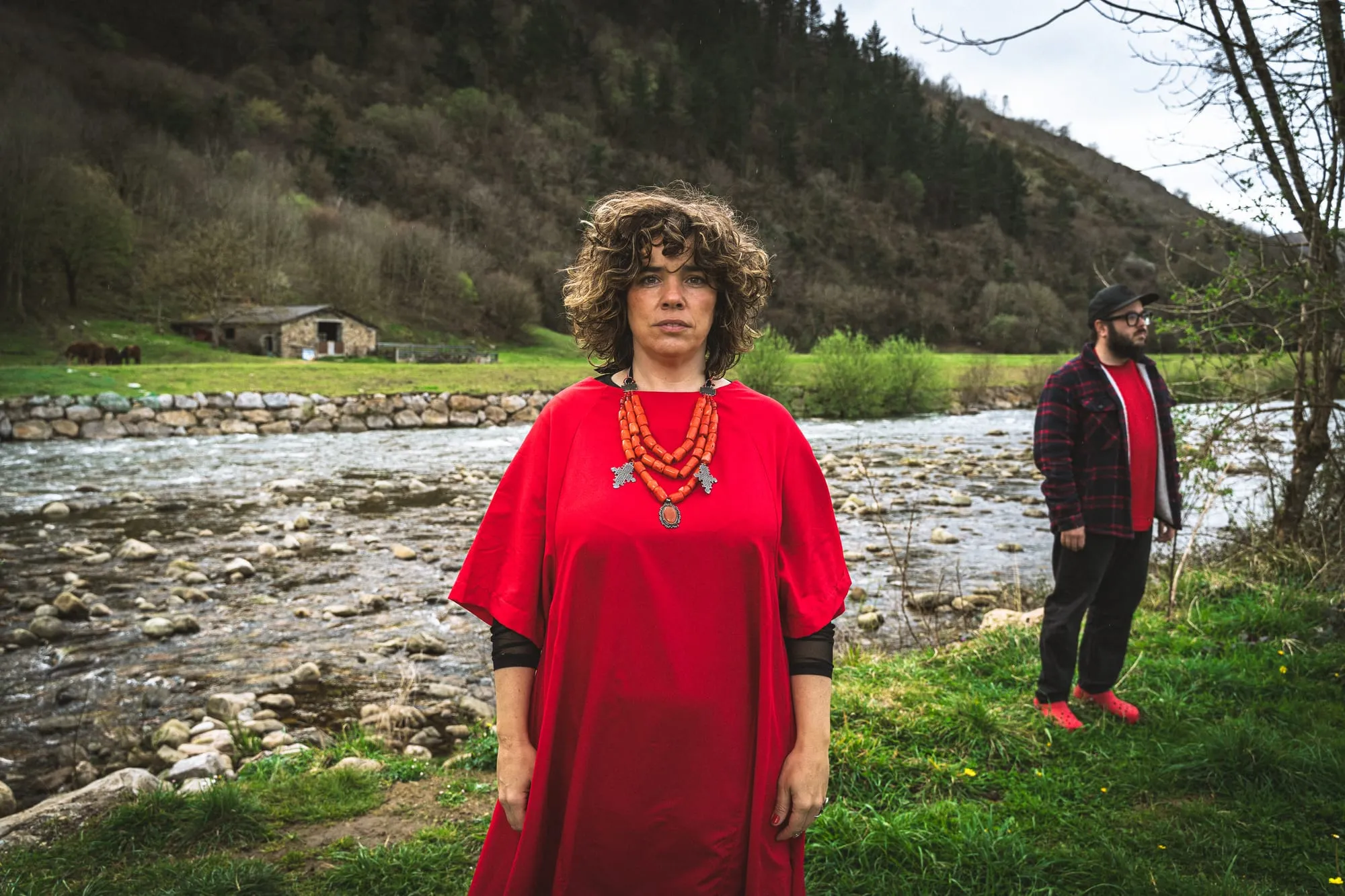La cantante de Casapalma, con túnica roja y collar de coral, mira a cámara junto a su compañero ante un río, una cabaña de piedra y un prado con ganado