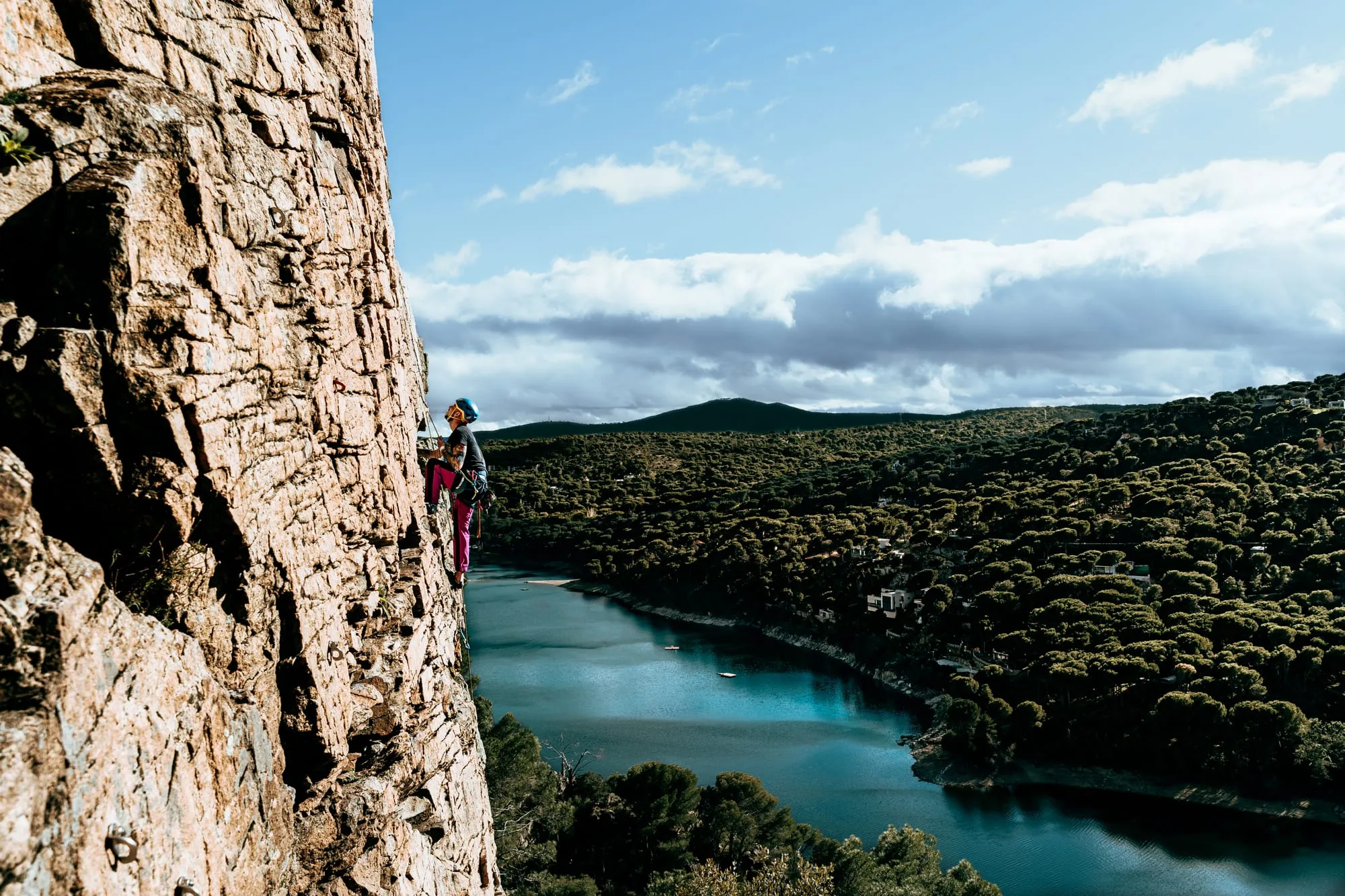 Escaladora con casco azul en una pared de granito sobre un embalse turquesa rodeado de pinares