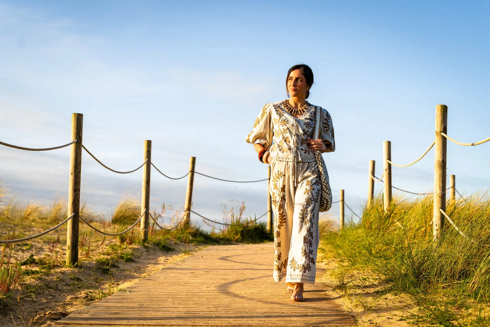 Modelo con un conjunto crema de bordados ocre caminando por la pasarela de madera de las dunas al atardecer