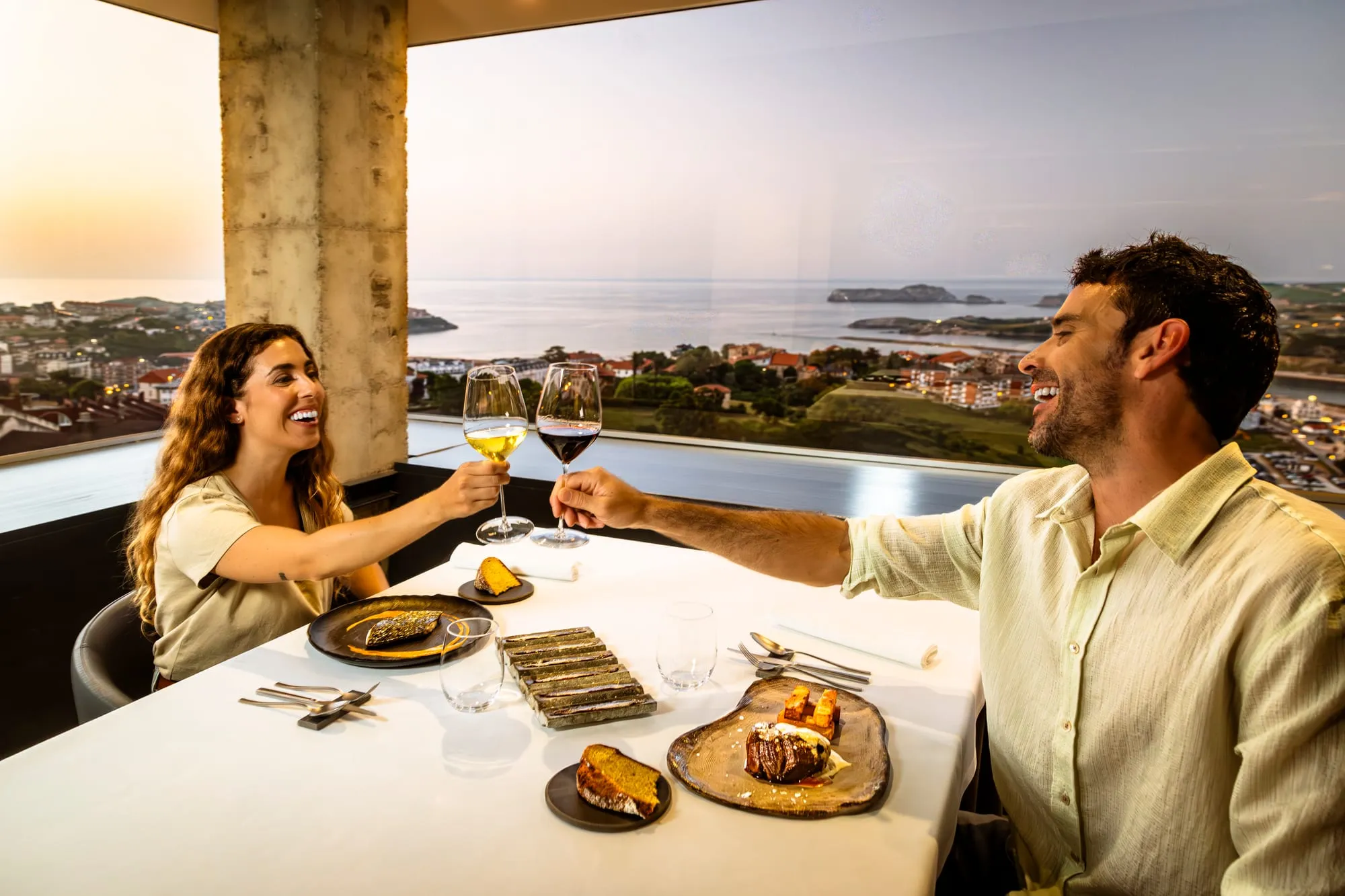 Pareja brindando con vino en un restaurante con vistas a la costa de Cantabria al atardecer