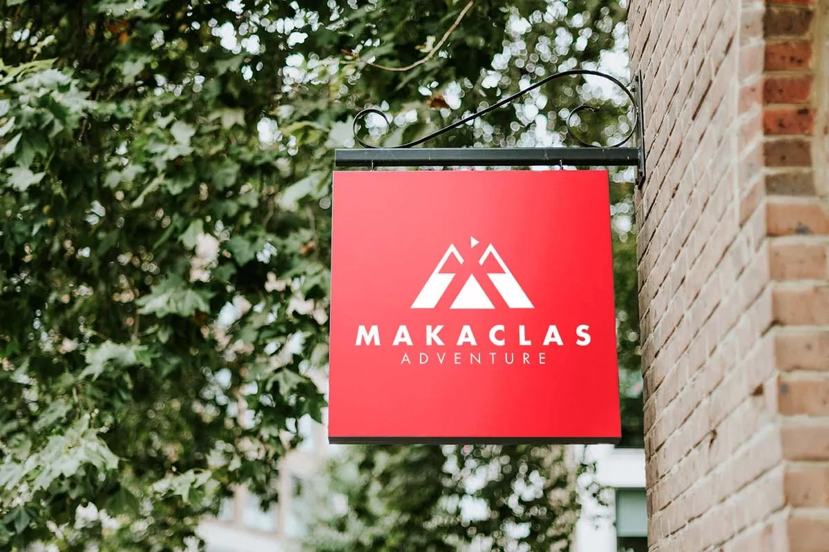 Rótulo rojo con el logotipo de Makaclas colgado en una fachada de ladrillo junto a un árbol