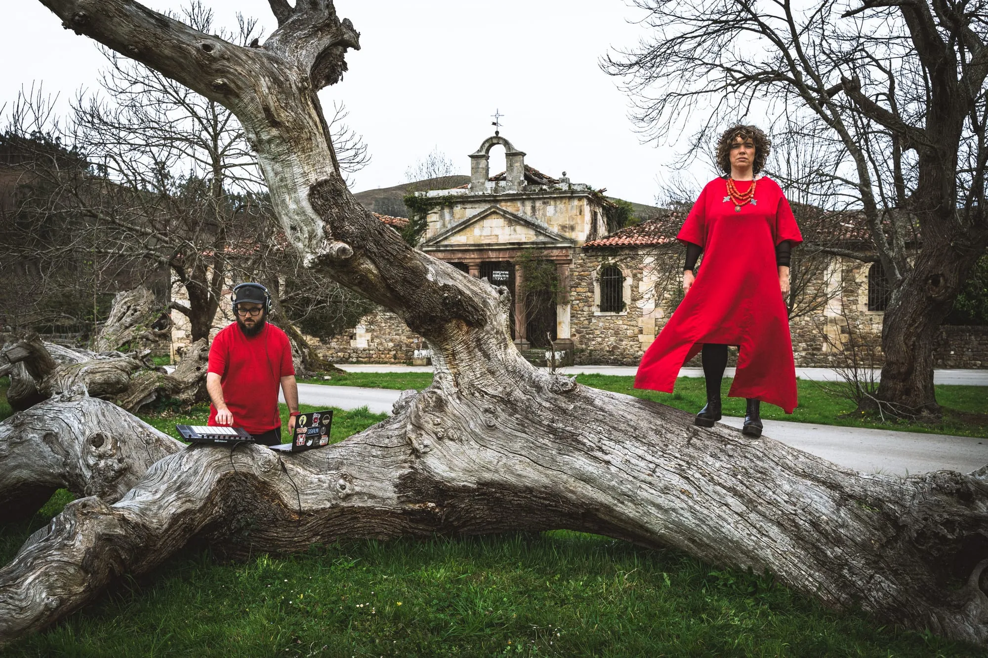 El dúo sobre un tronco caído frente a una ermita de piedra, él con el portátil y ella de rojo