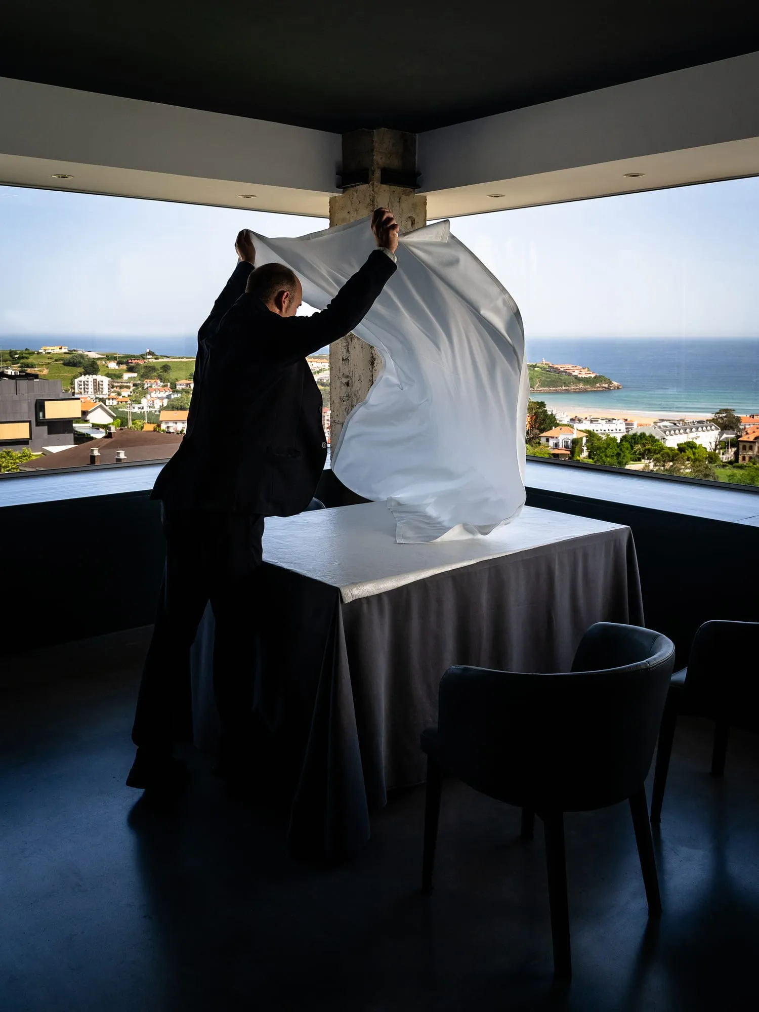 Un camarero de Emma extiende el mantel sobre una mesa frente al ventanal con vistas a la playa de Suances