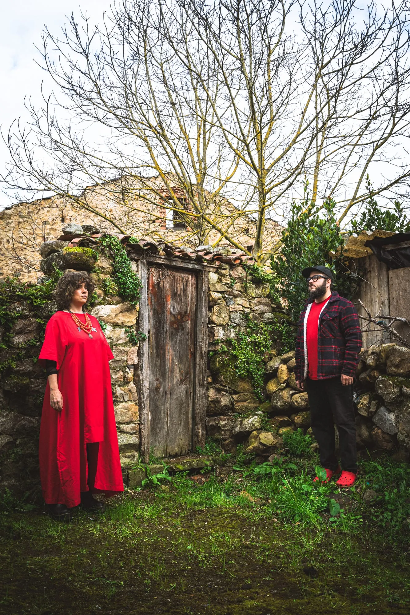 Los dos músicos ante la puerta de madera de una casa de piedra en ruinas, ella de rojo