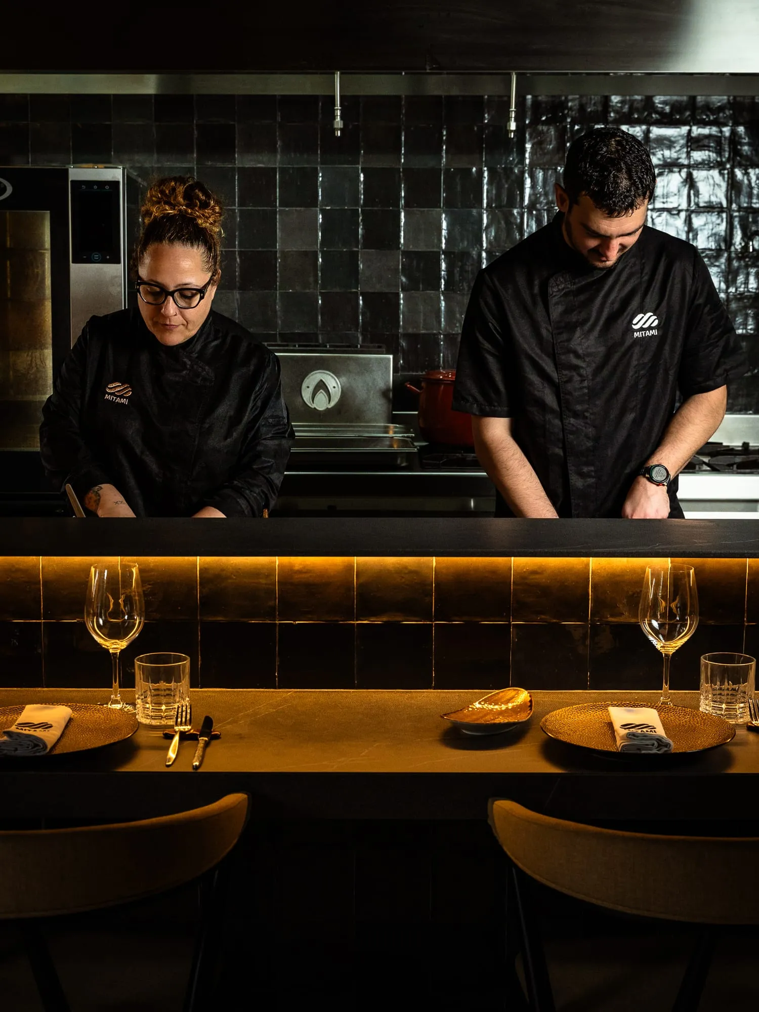 Dos cocineros con chaquetilla negra trabajando tras la barra de la cocina abierta de azulejo verde