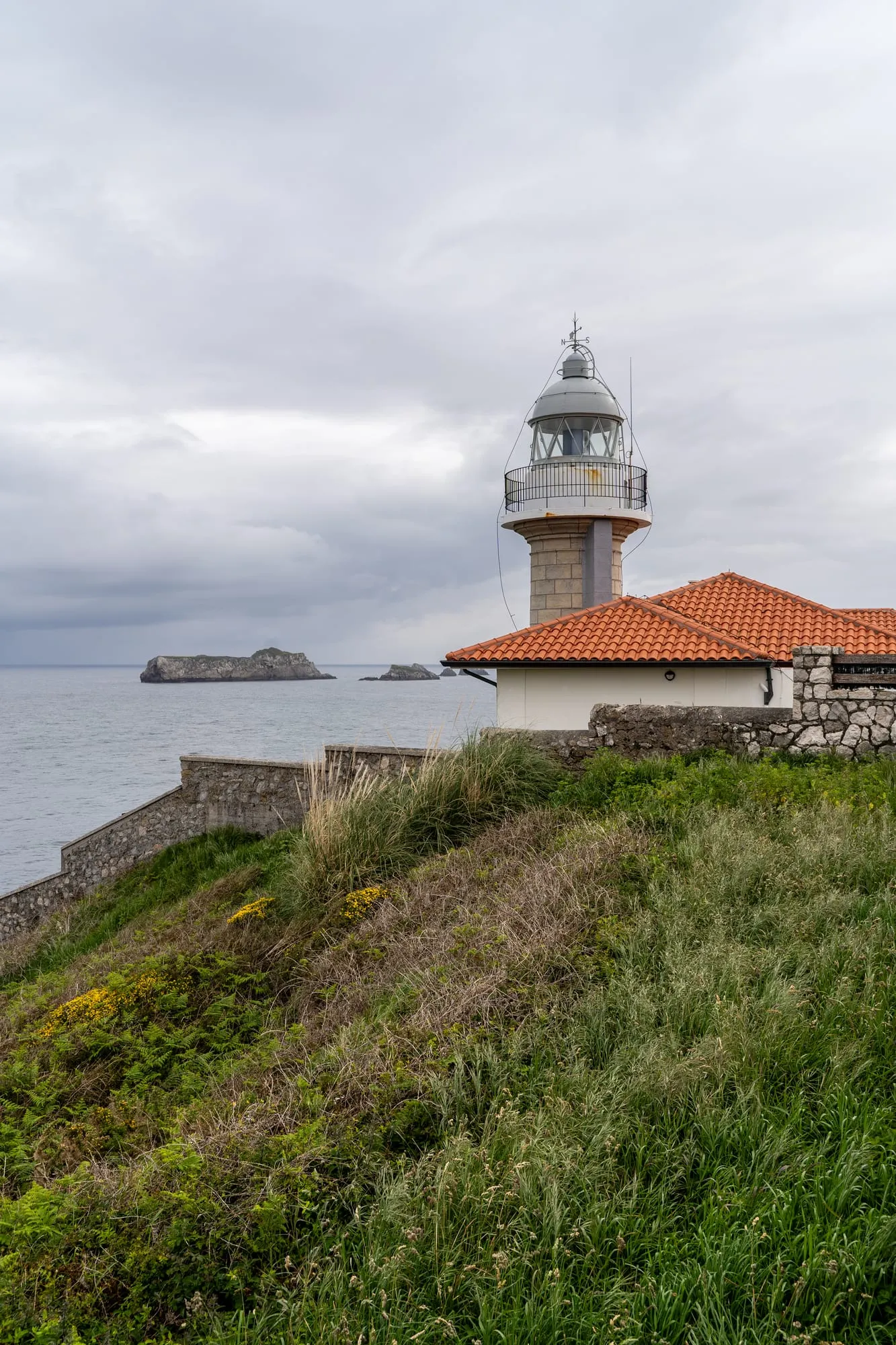 Faro con tejado rojo sobre un prado verde y un islote rocoso al fondo en el Cantábrico