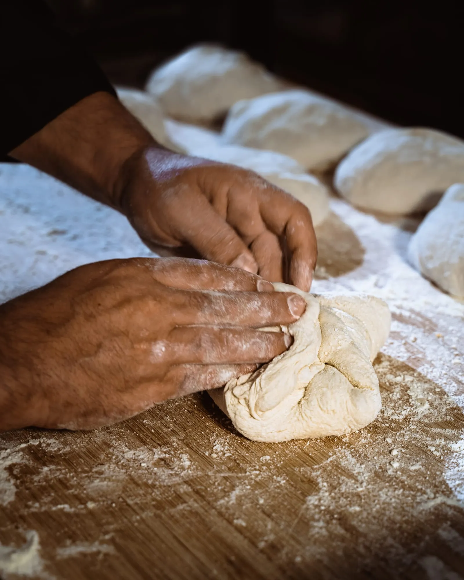 Manos enharinadas plegando una pieza de masa con bollos fermentando al fondo
