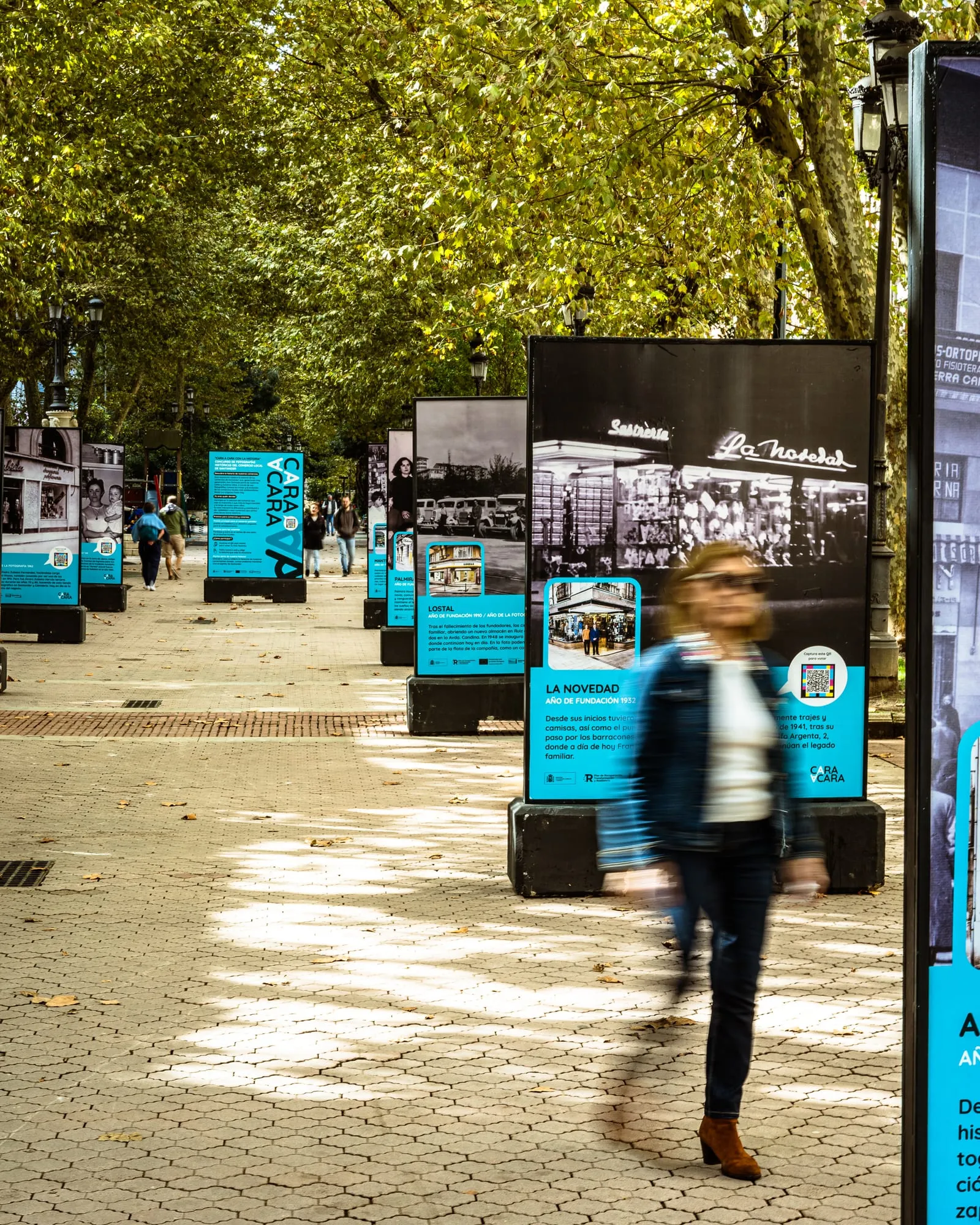 Paseo arbolado con la hilera de paneles de la exposición de comercios históricos y una mujer caminando