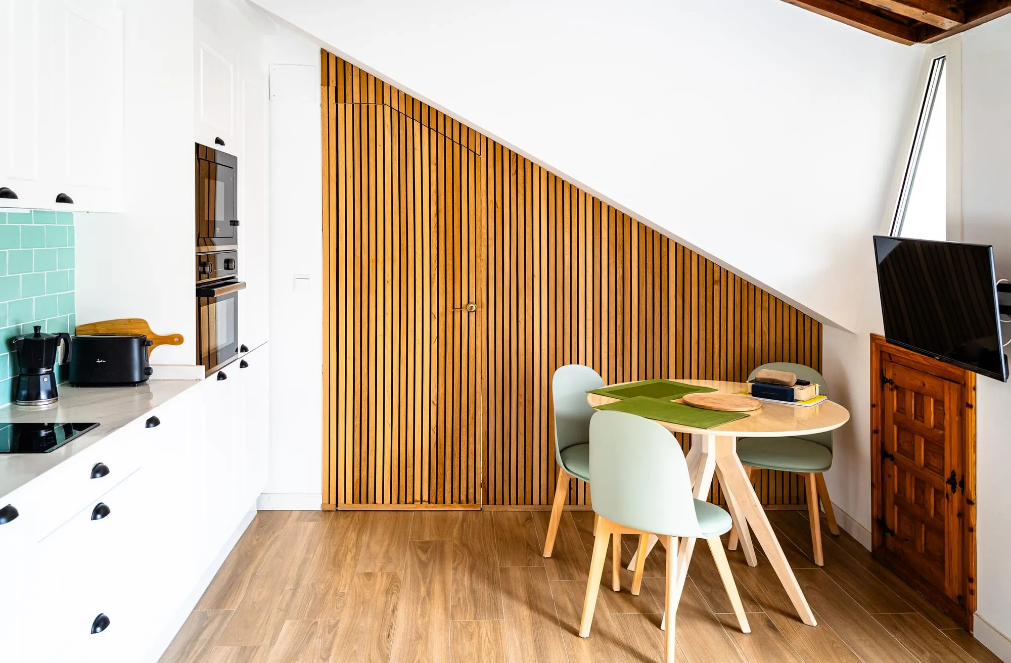 Cocina y comedor luminosos con mesa redonda y una pared de listones de madera bajo la escalera
