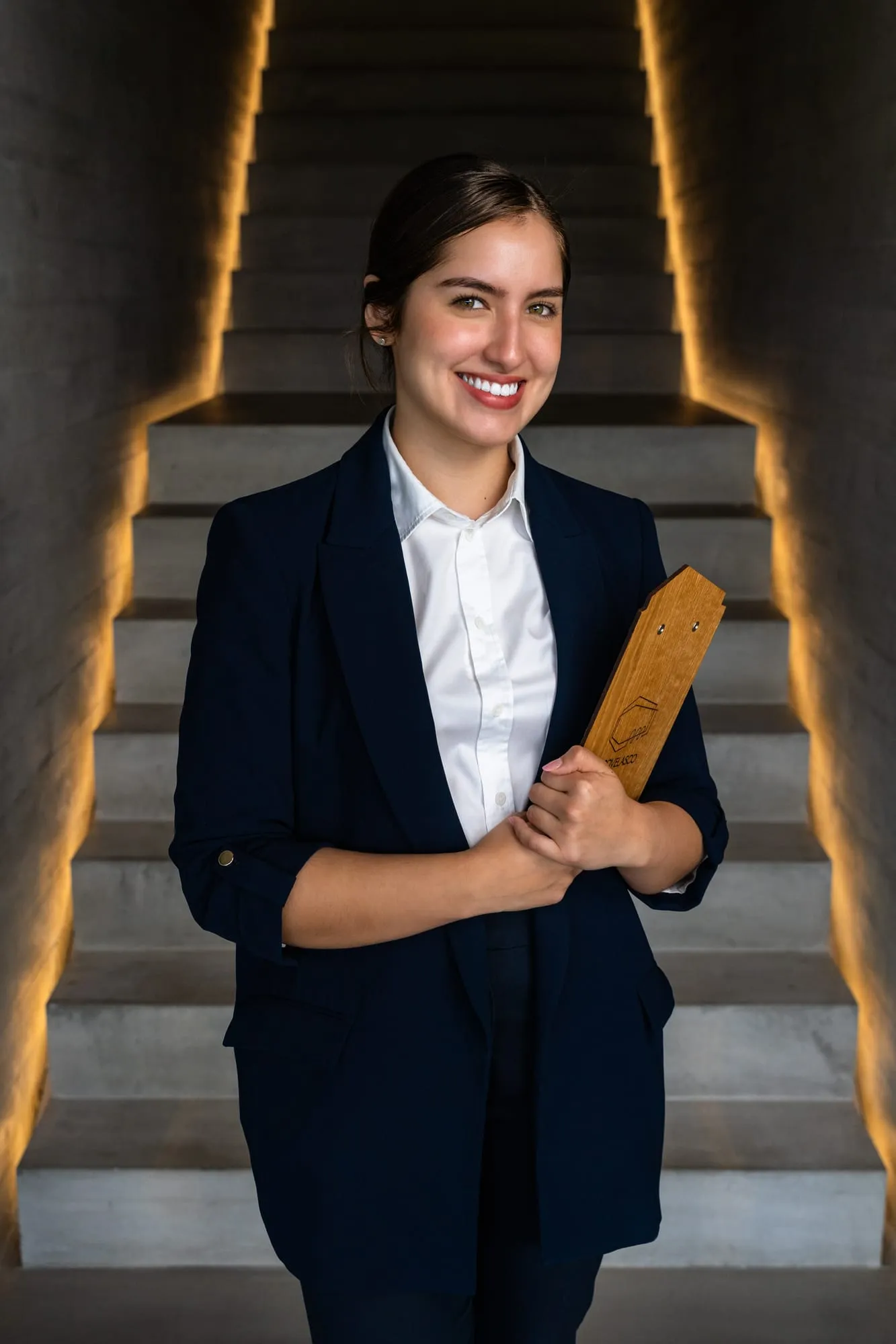 Miembro del equipo de sala, con americana azul marino y carta de madera, en la escalera iluminada del hotel