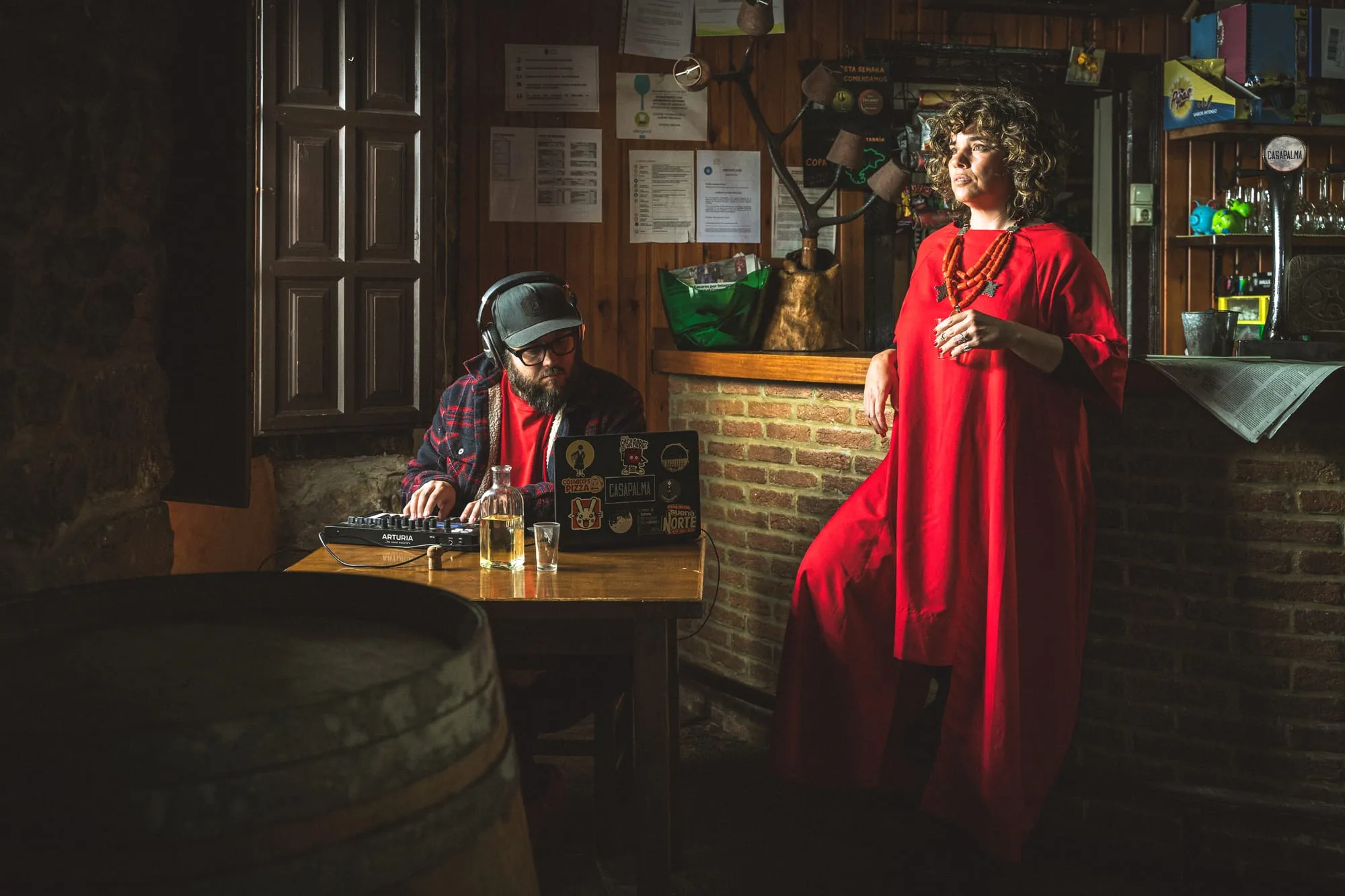 Él con teclado y portátil en la mesa de una taberna; ella, de rojo, de pie junto a la barra