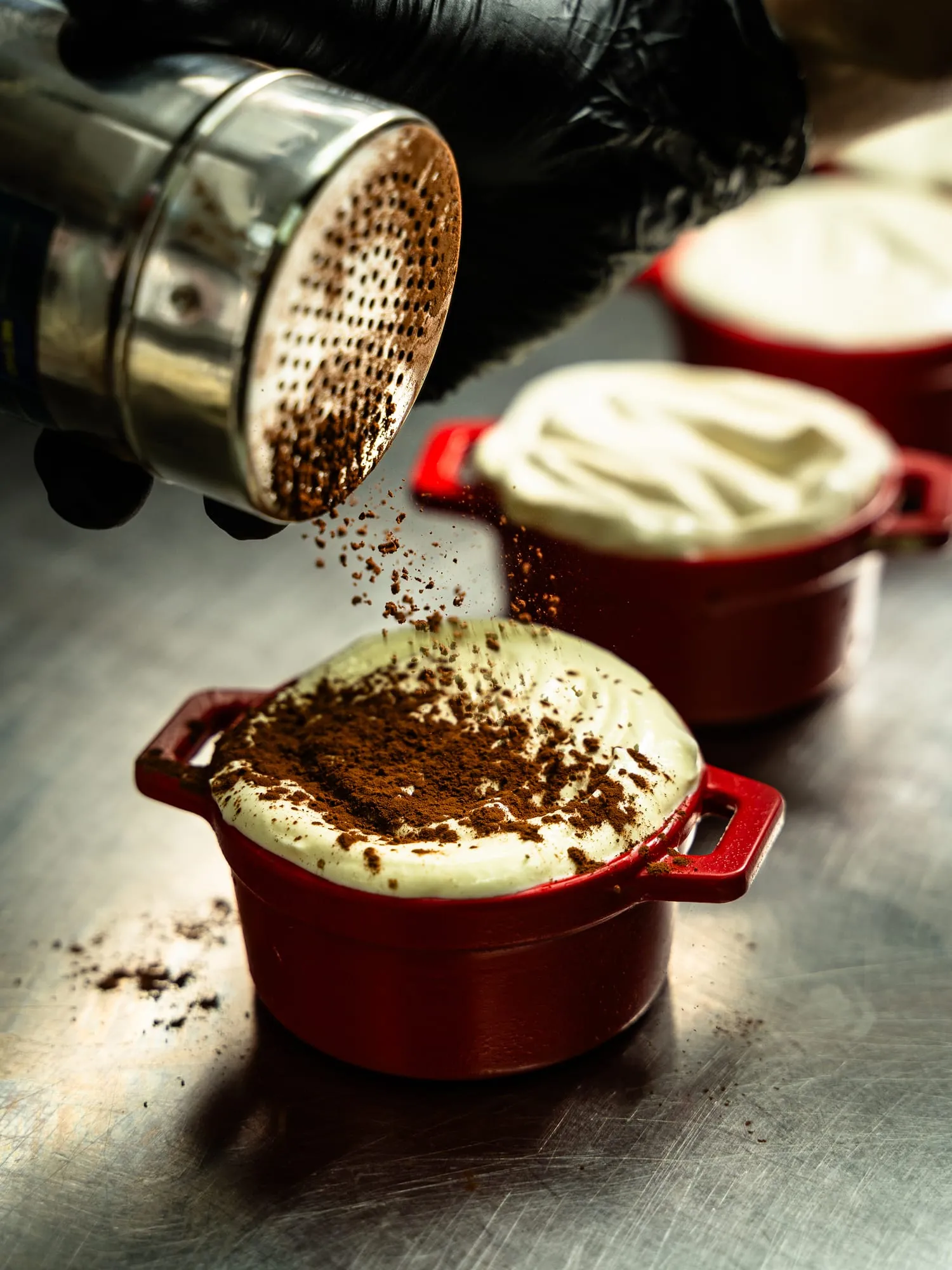 Mano espolvoreando cacao sobre un tiramisú servido en una mini cocotte roja
