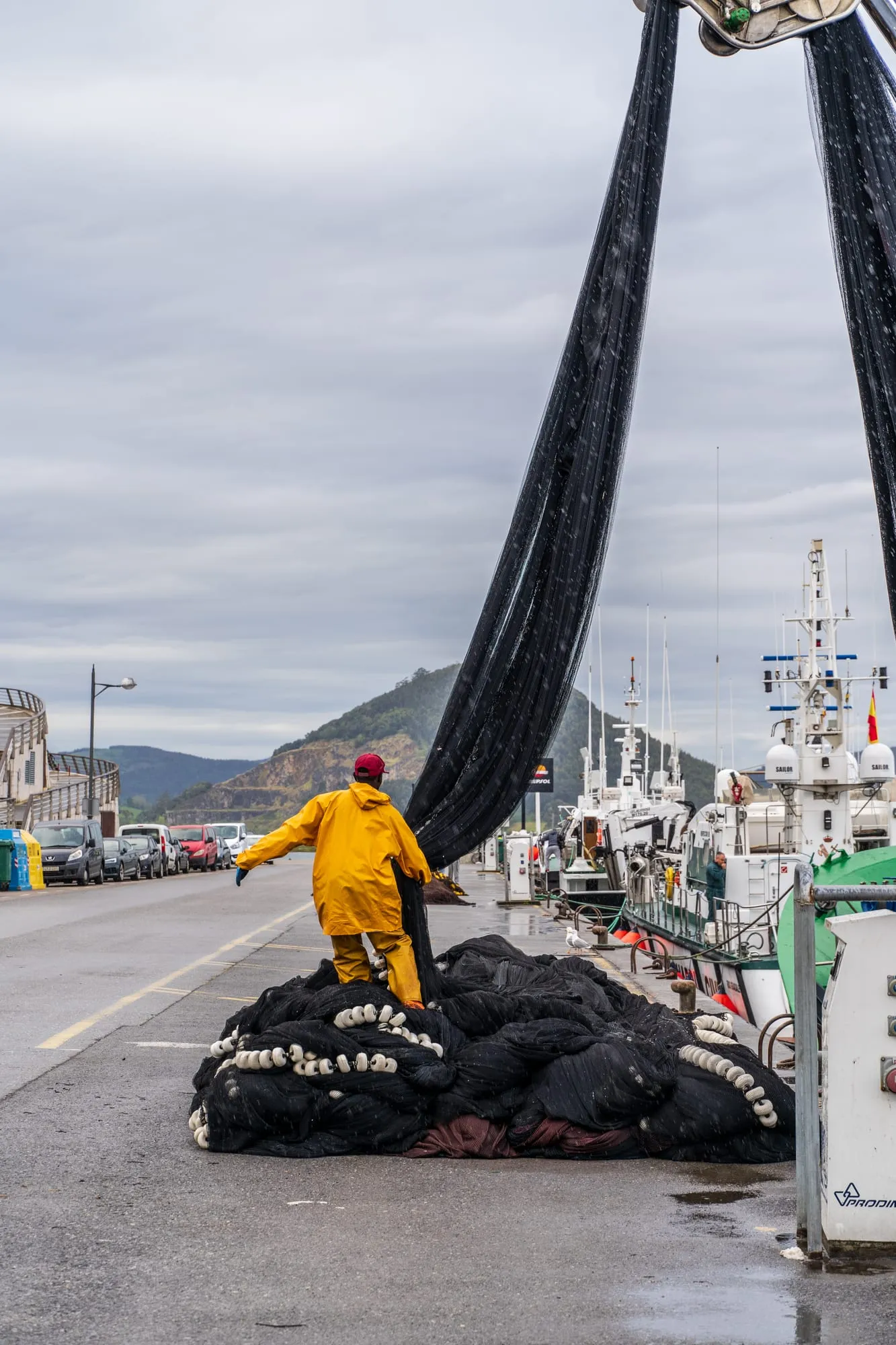 Pescador con traje de aguas amarillo sobre un montón de redes en el muelle, con barcos amarrados
