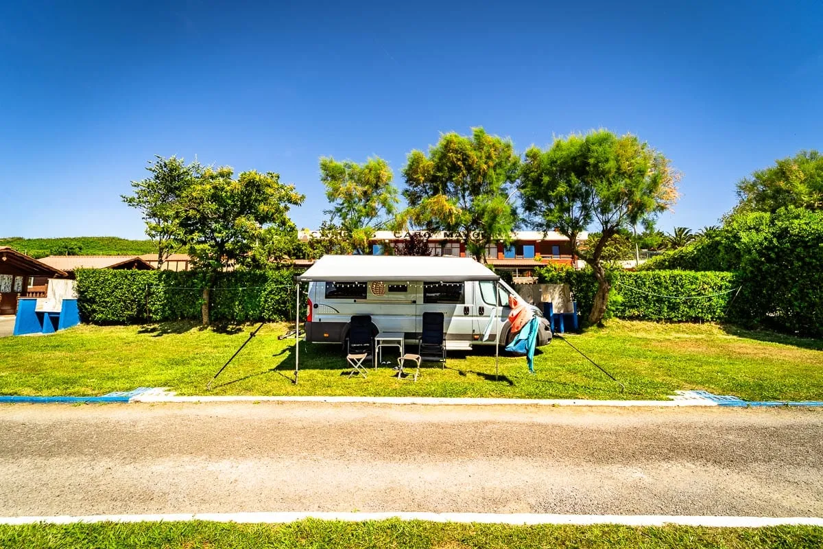 Parcela de acampada con una autocaravana y el toldo abierto entre setos, bajo el cielo azul