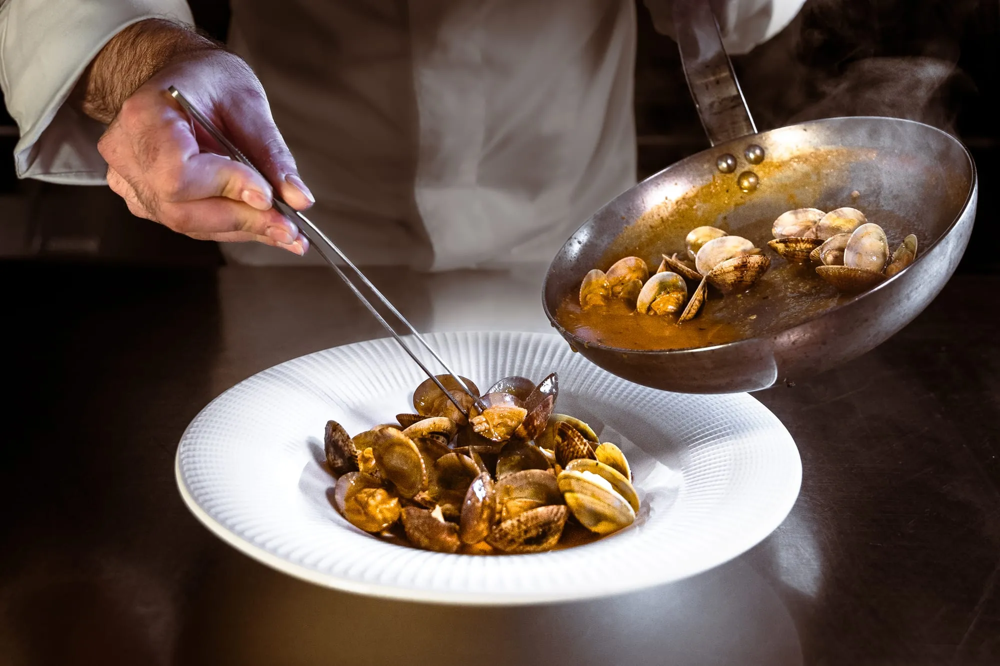 El chef emplata almejas a la marinera desde la sartén en un plato hondo blanco