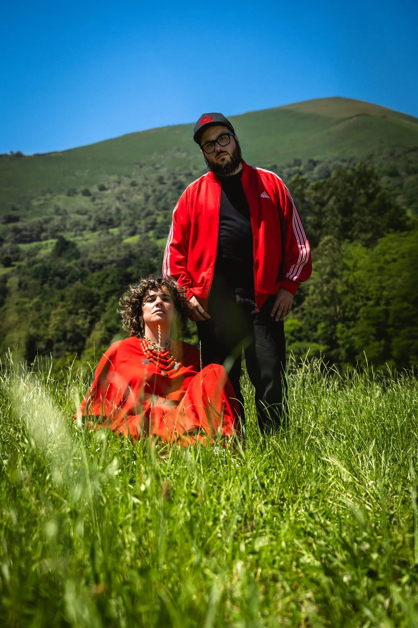 Él de pie y ella sentada entre la hierba de un prado de montaña, ambos de rojo