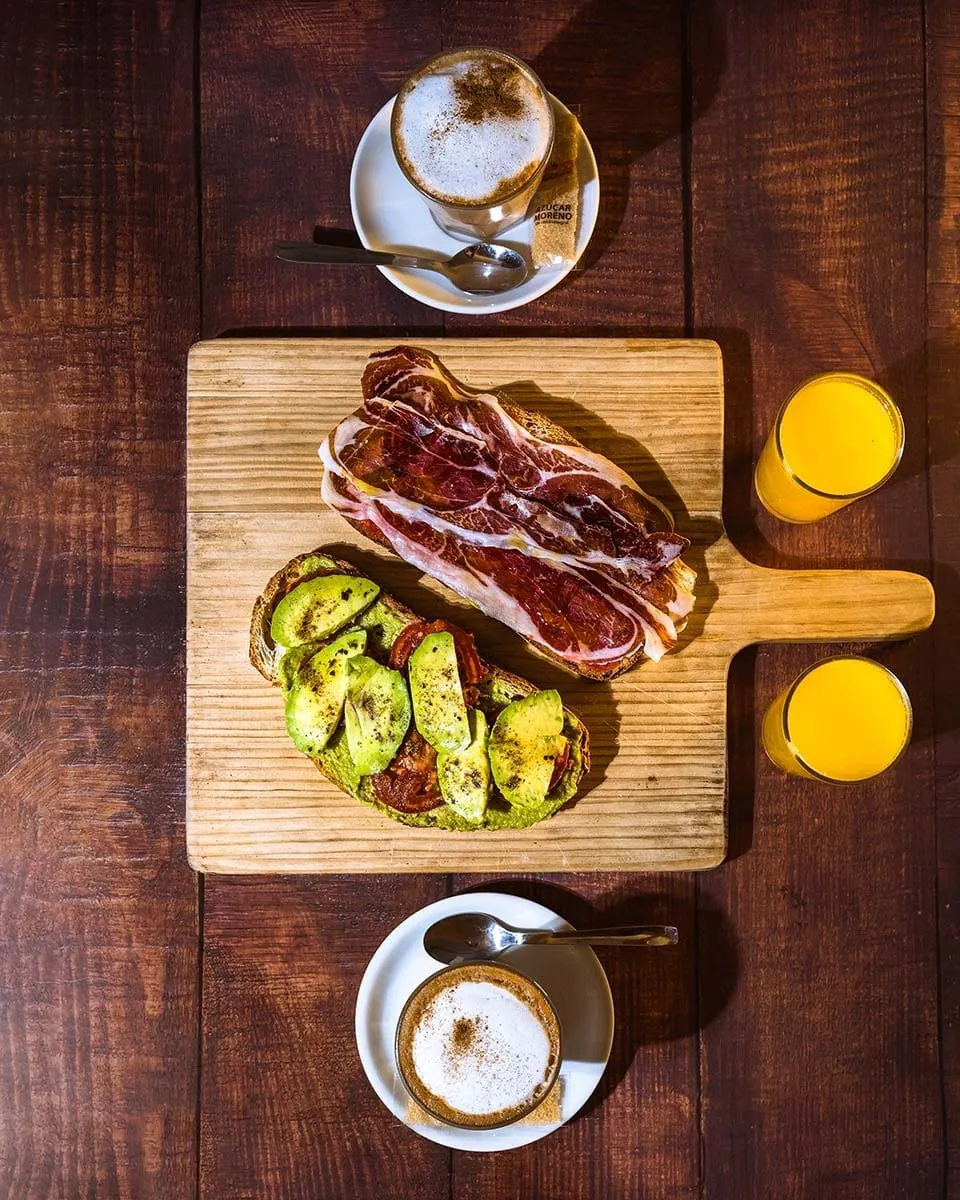 Tabla de desayuno vista desde arriba con tostadas de jamón y aguacate, dos cafés y dos zumos