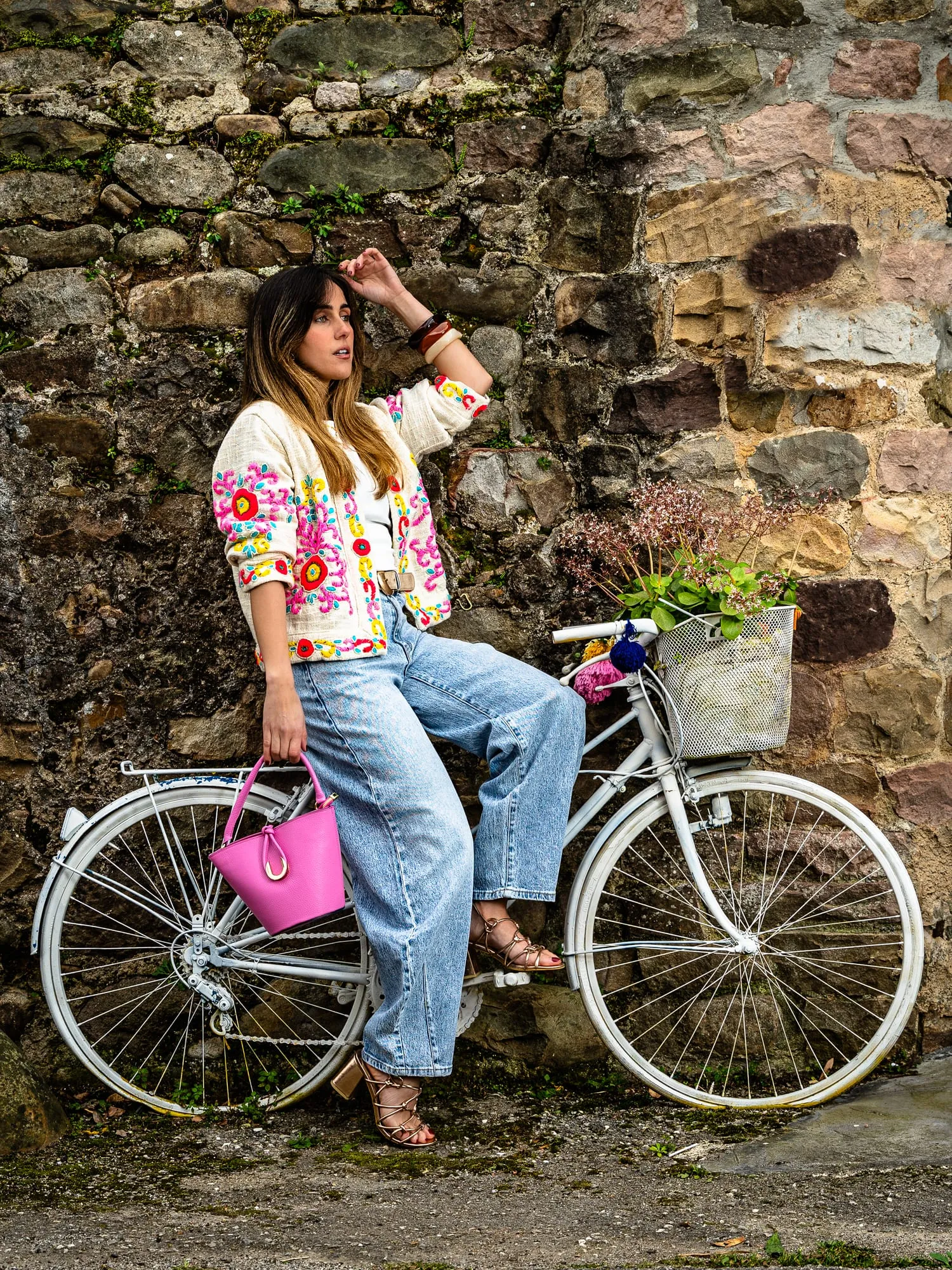 Modelo con chaqueta de flores, vaqueros y bolso rosa junto a una bicicleta blanca ante un muro de piedra