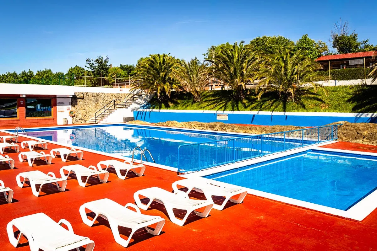 Piscina del camping con tumbonas blancas sobre el solárium rojo y palmeras al fondo