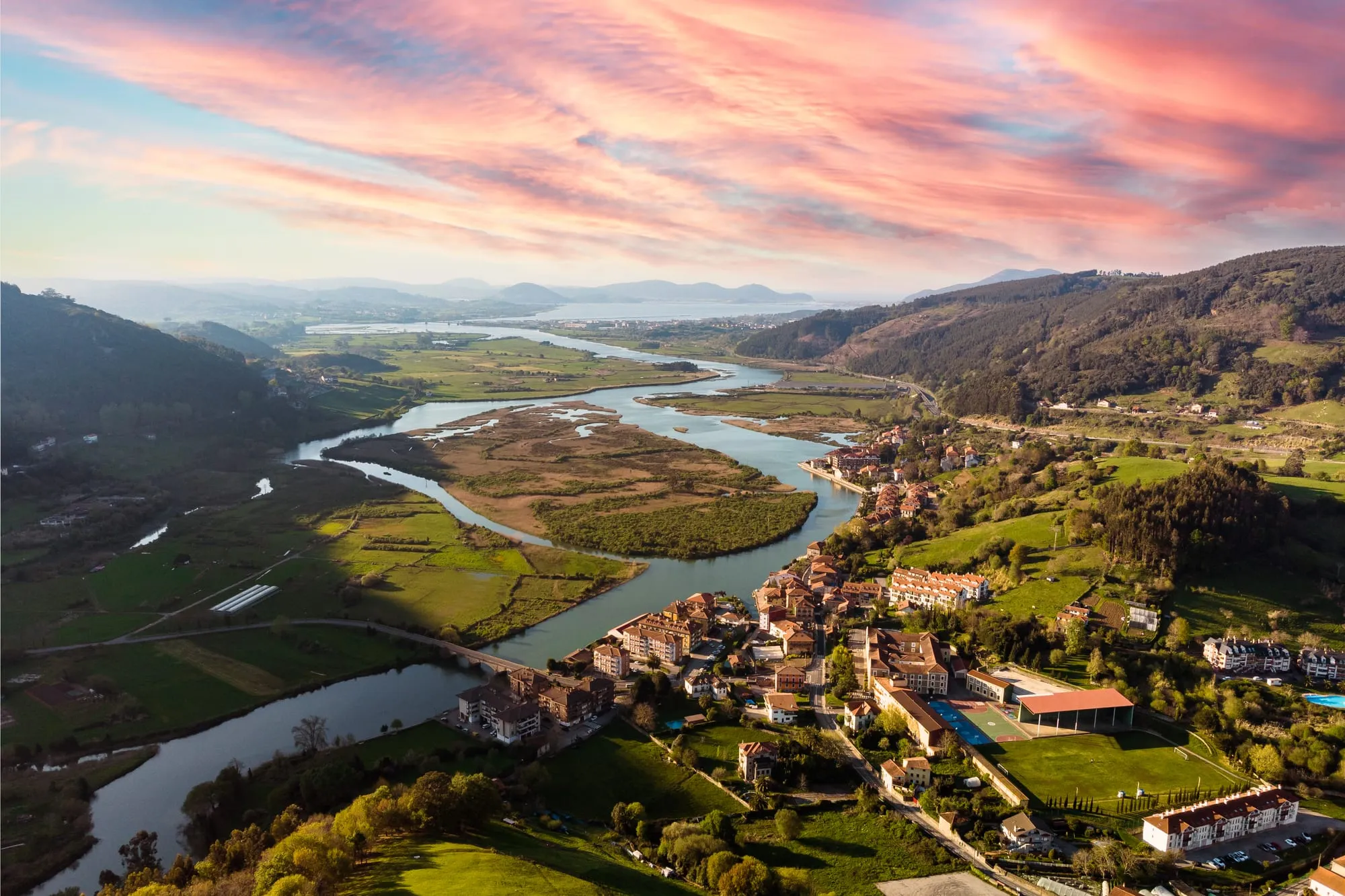 La ría serpentea entre marismas junto al casco de Limpias bajo un cielo rosa, vista aérea