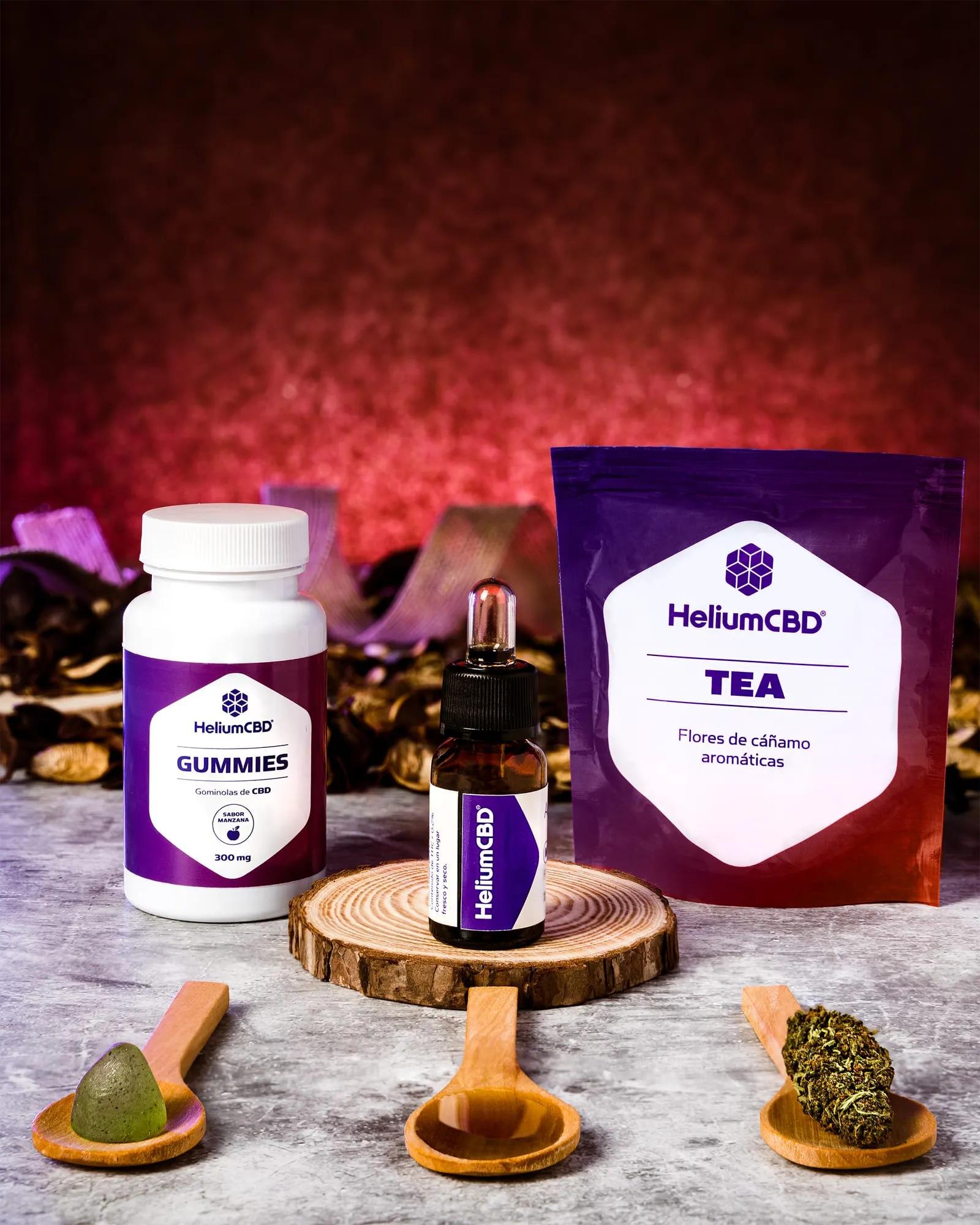 Bote de gominolas, frasco de aceite y bolsa de té HeliumCBD con tres cucharas de madera sobre mármol y fondo rojo