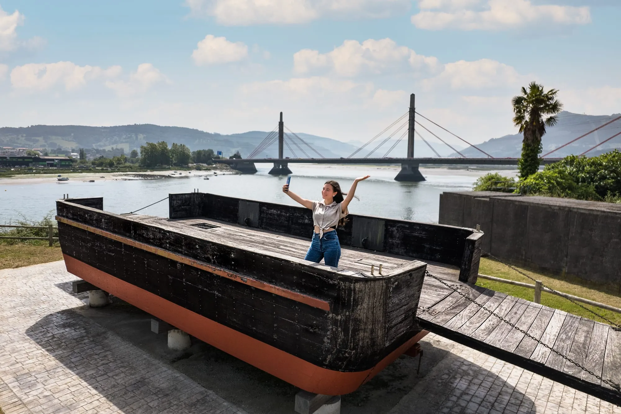 Una joven se hace un selfi sobre una barcaza de madera, con el puente de la ría al fondo