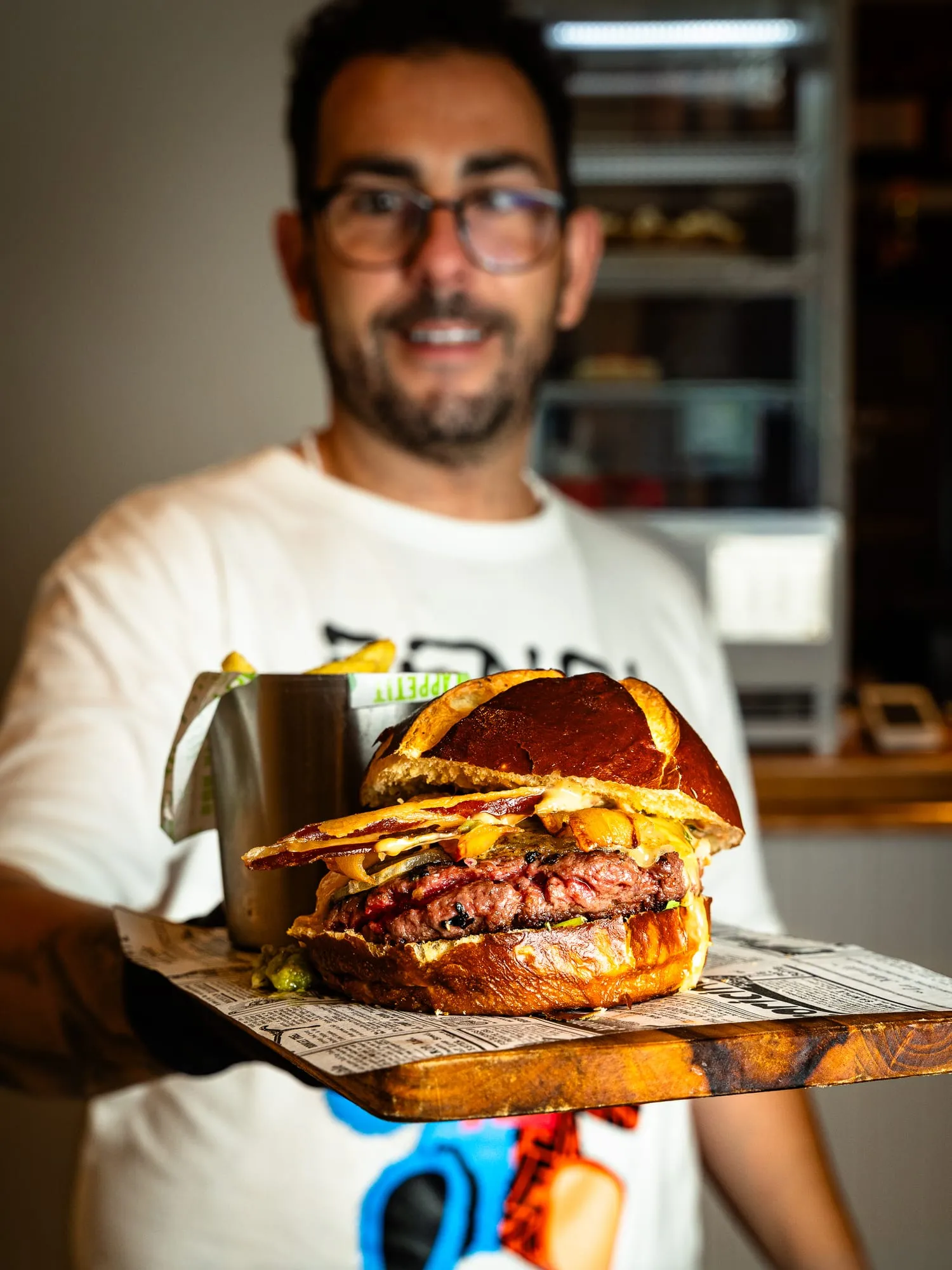 Cocinero sonriente de The Duke's enseña a cámara una hamburguesa con patatas sobre una tabla de madera