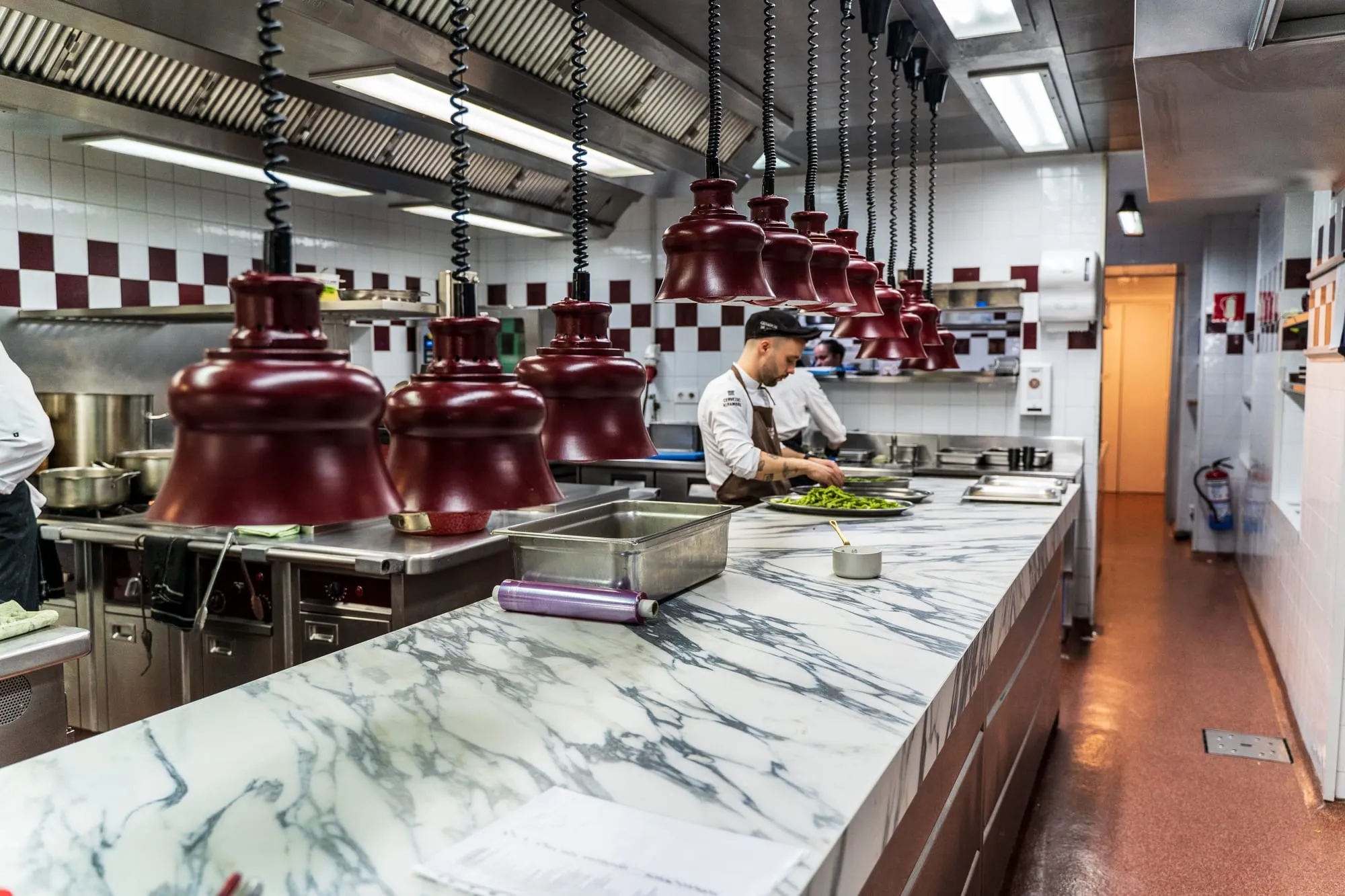 Cocina del Cenador de Amós con pase de mármol, lámparas granate y cocineros emplatando