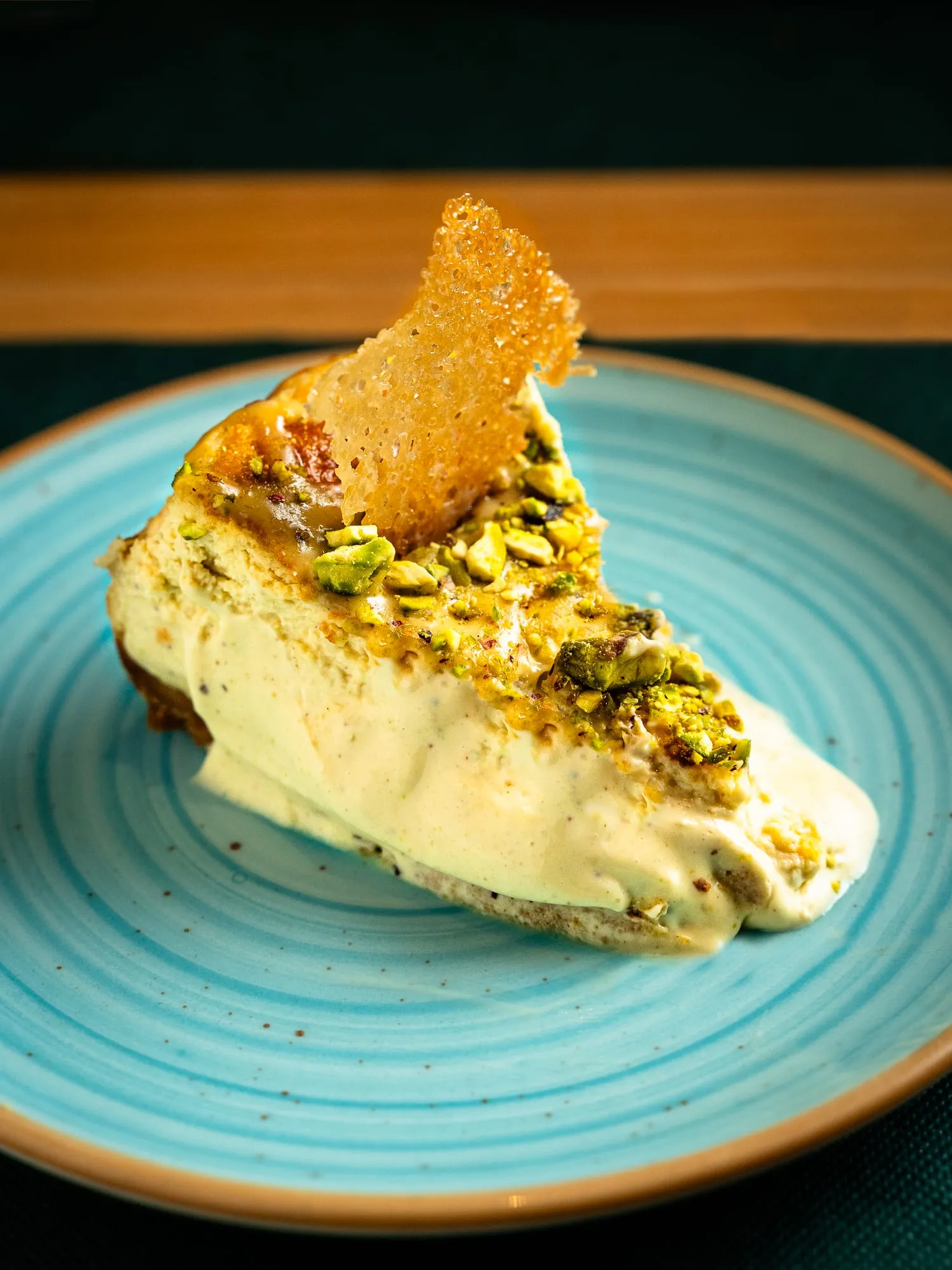 Porción de tarta de queso con pistacho y teja crujiente en plato turquesa
