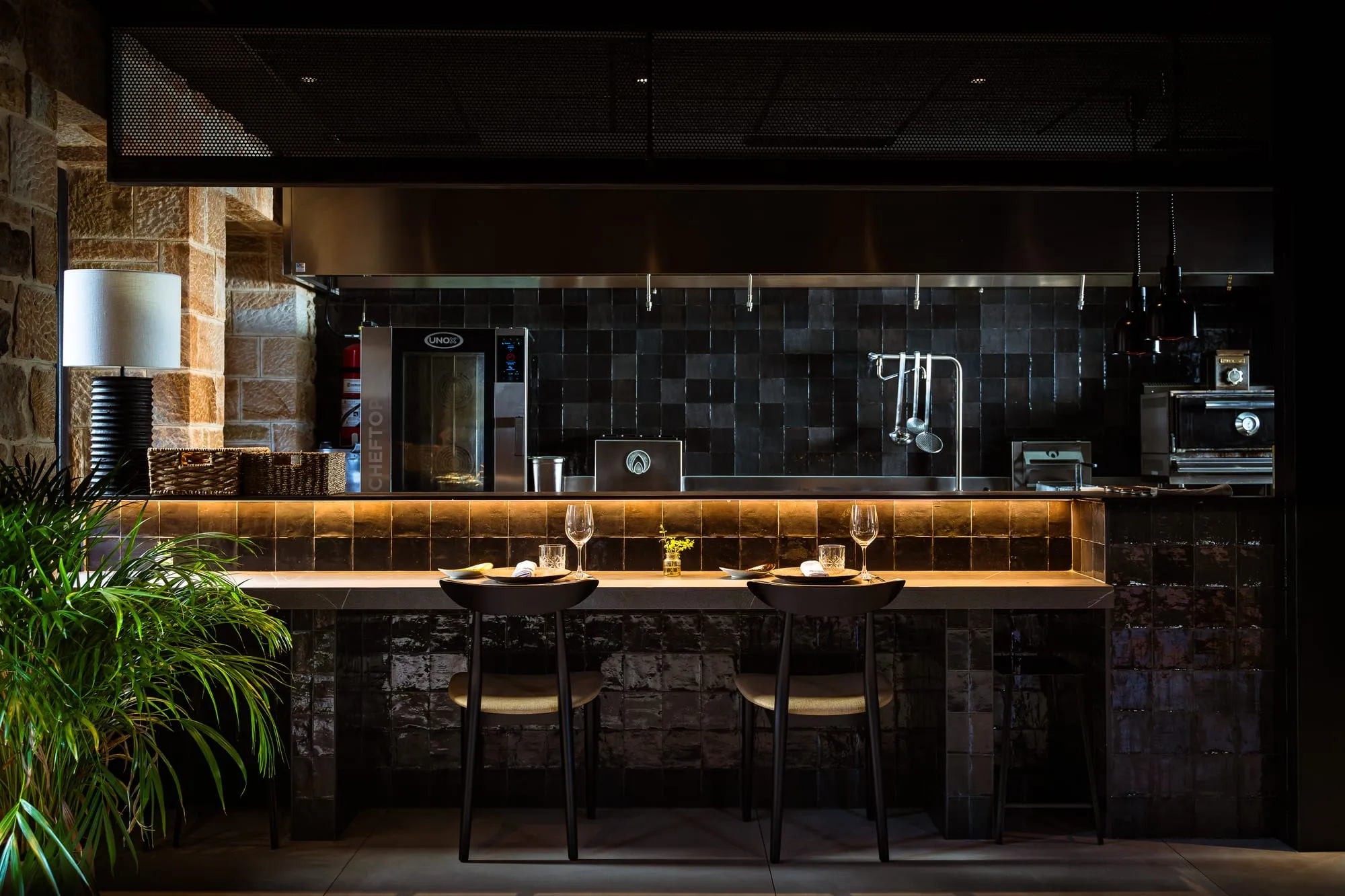 Barra y cocina abierta de Mitami de noche, con lámpara cálida, azulejo verde y plantas