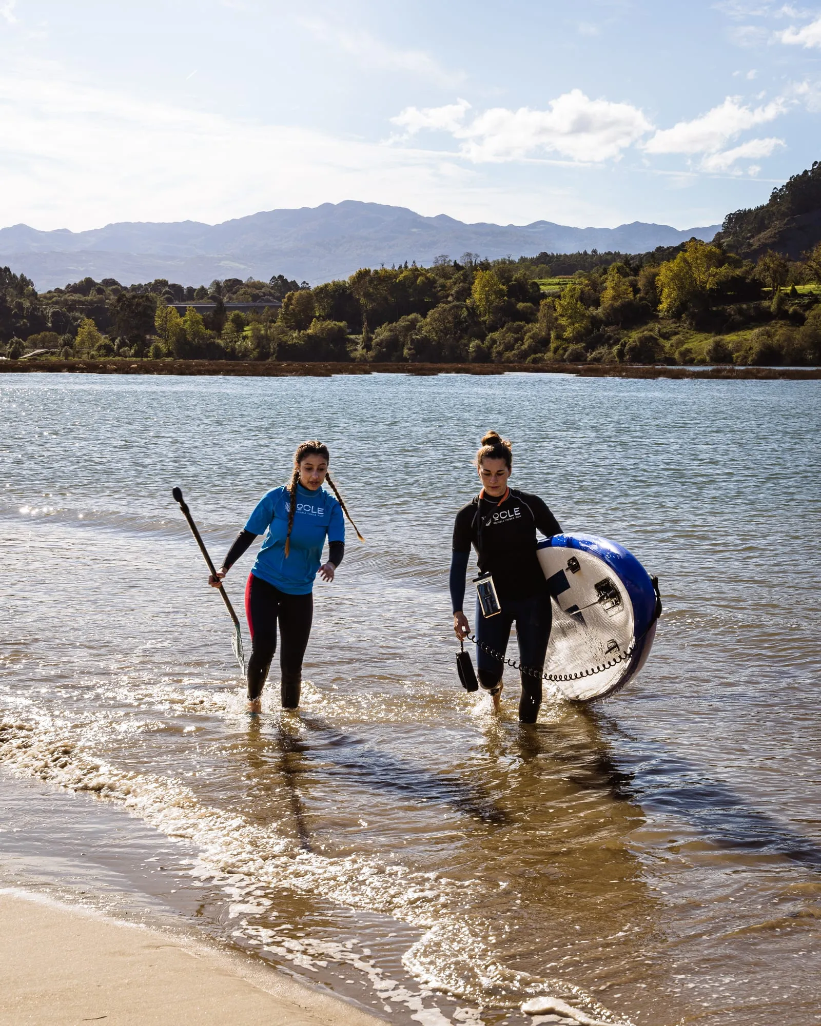 Dos chicas salen del agua sonriendo con la tabla bajo el brazo y el remo tras la travesía
