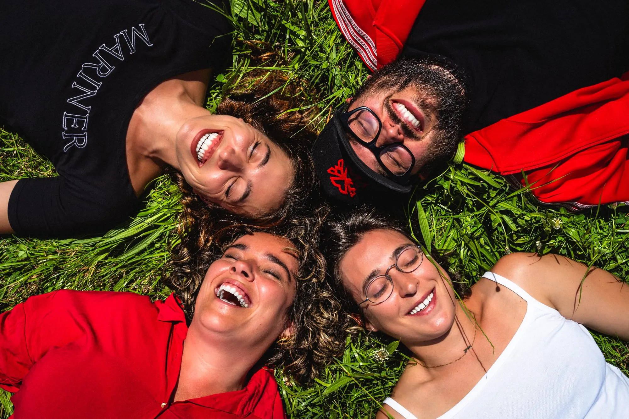 Cuatro músicos tumbados en la hierba con las cabezas juntas, riendo, vistos desde arriba