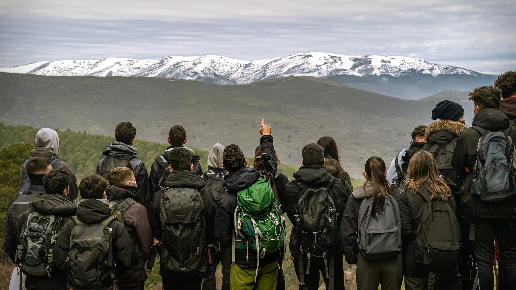 Grupo de senderistas de espaldas contemplando las cumbres nevadas de la sierra, uno señala con el brazo en alto