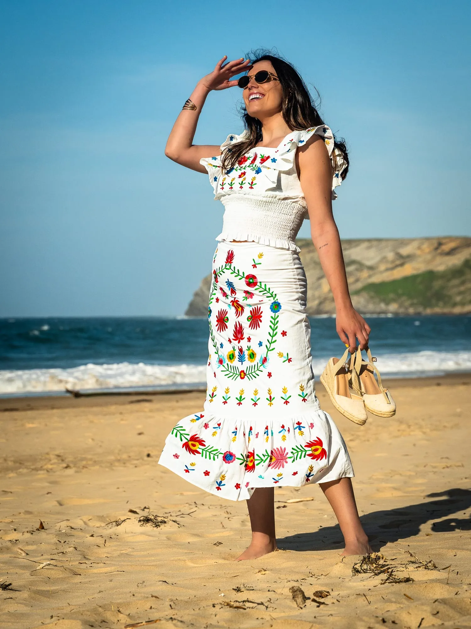 Modelo con vestido blanco de bordados de colores y las sandalias en la mano, en la playa con acantilados