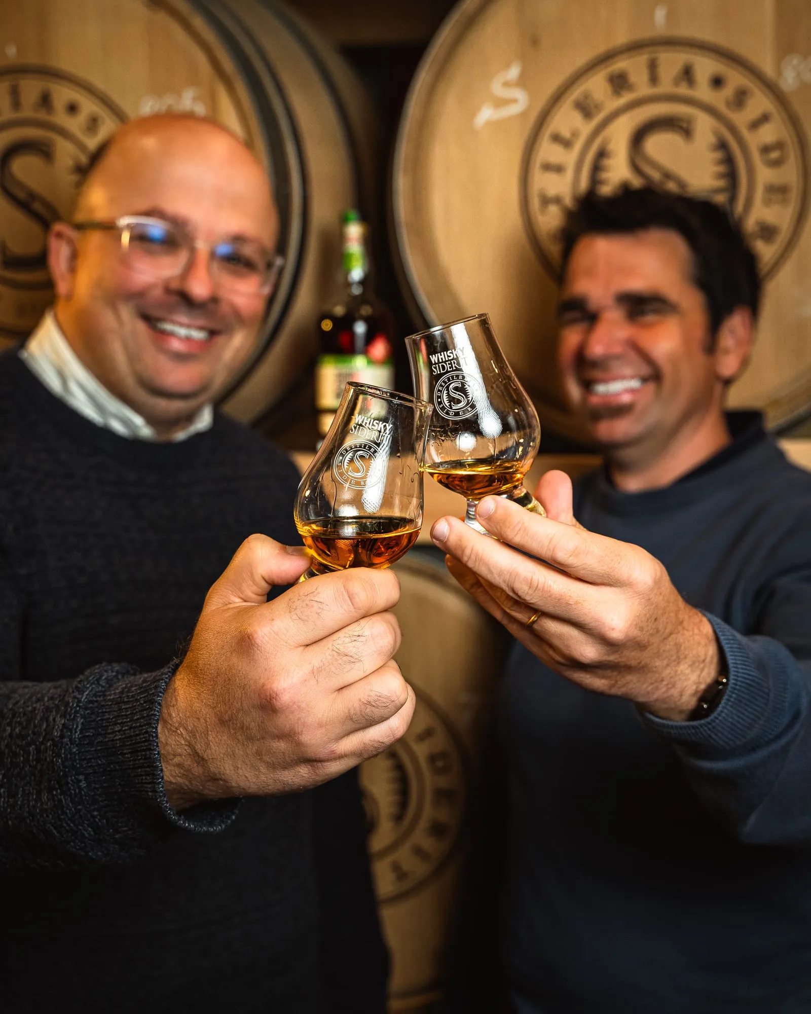 Los dos fundadores brindan con copas de whisky delante de las barricas de la destilería