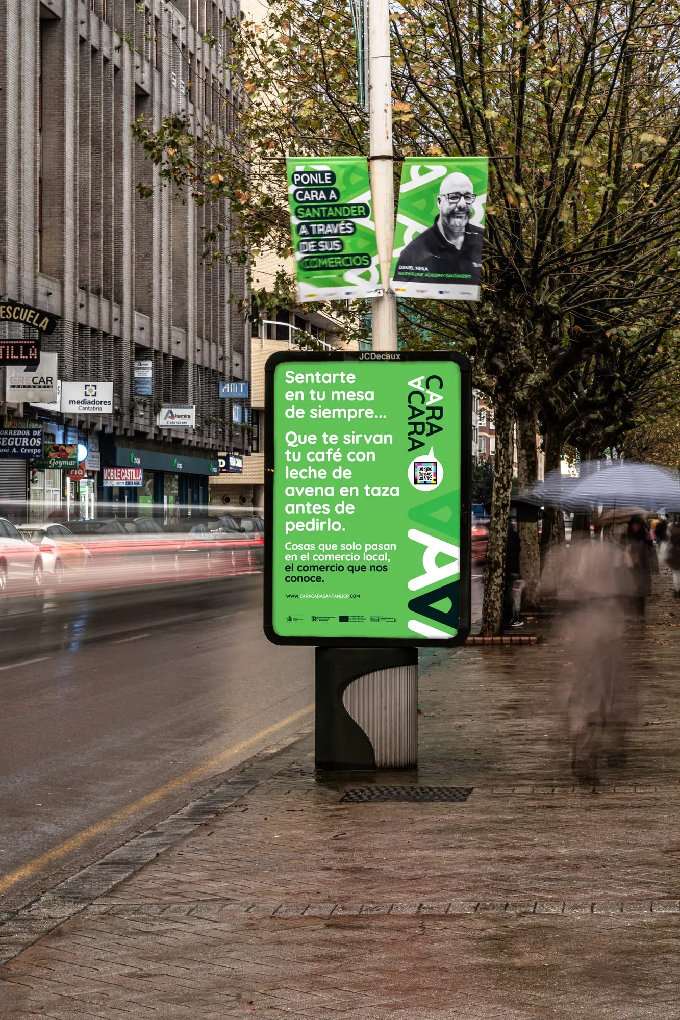 Mupi verde «Sentarte en tu mesa de siempre…» bajo dos banderolas de la campaña en una calle de Santander