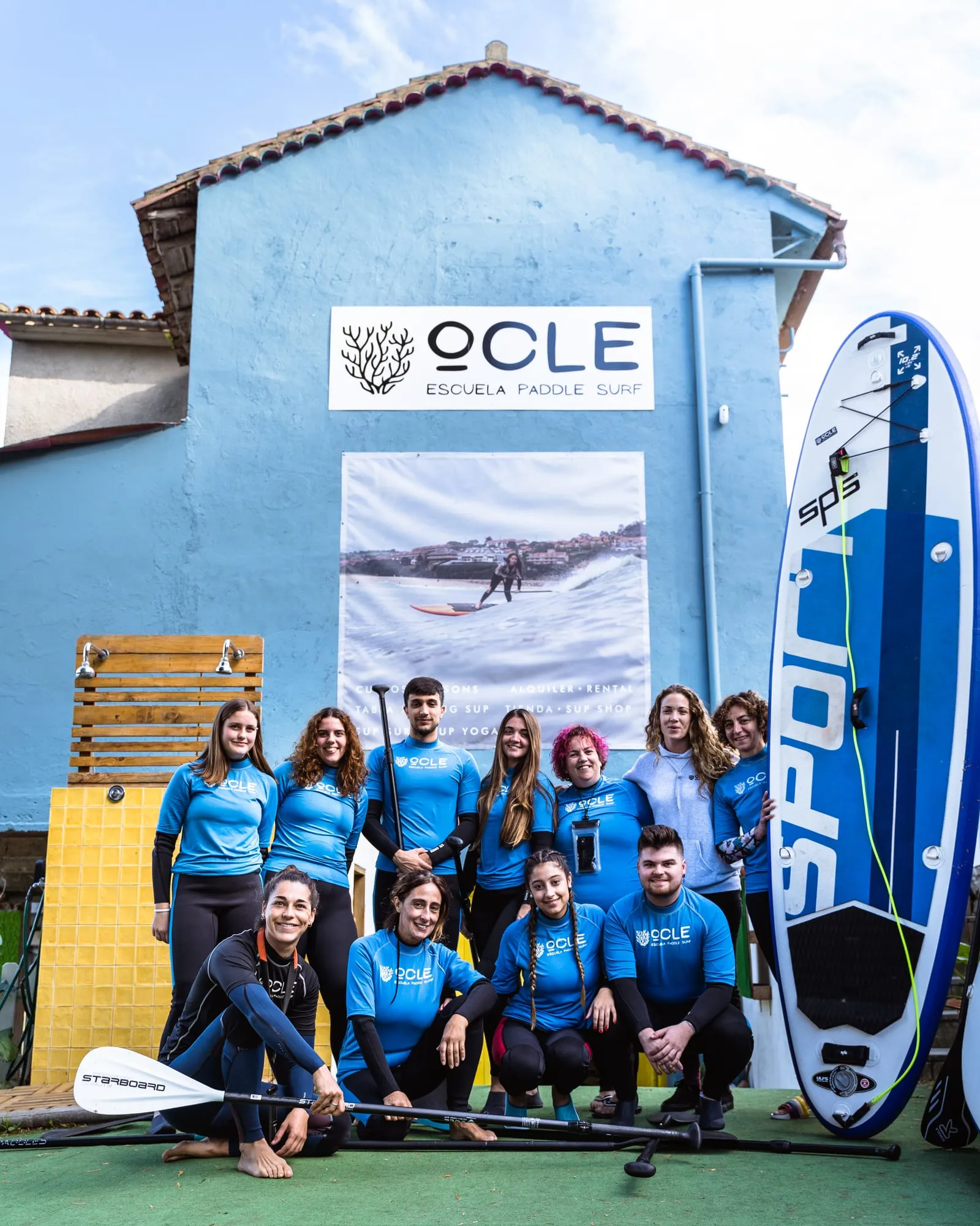 El equipo de Ocle con las licras azules, remos y tablas, posando ante la fachada de la escuela