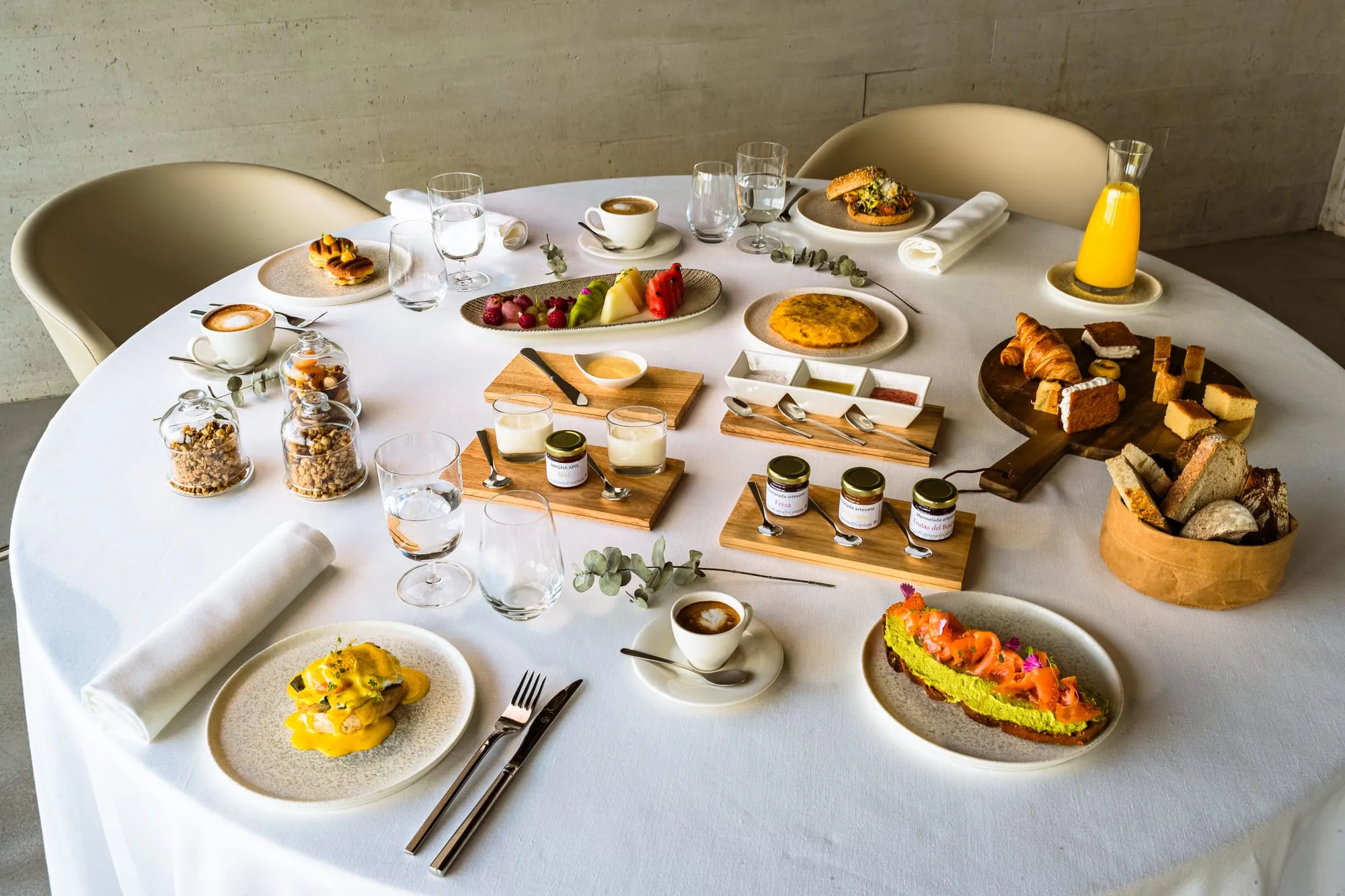 Desayuno completo servido sobre mantel blanco con zumo, bollería y platos salados
