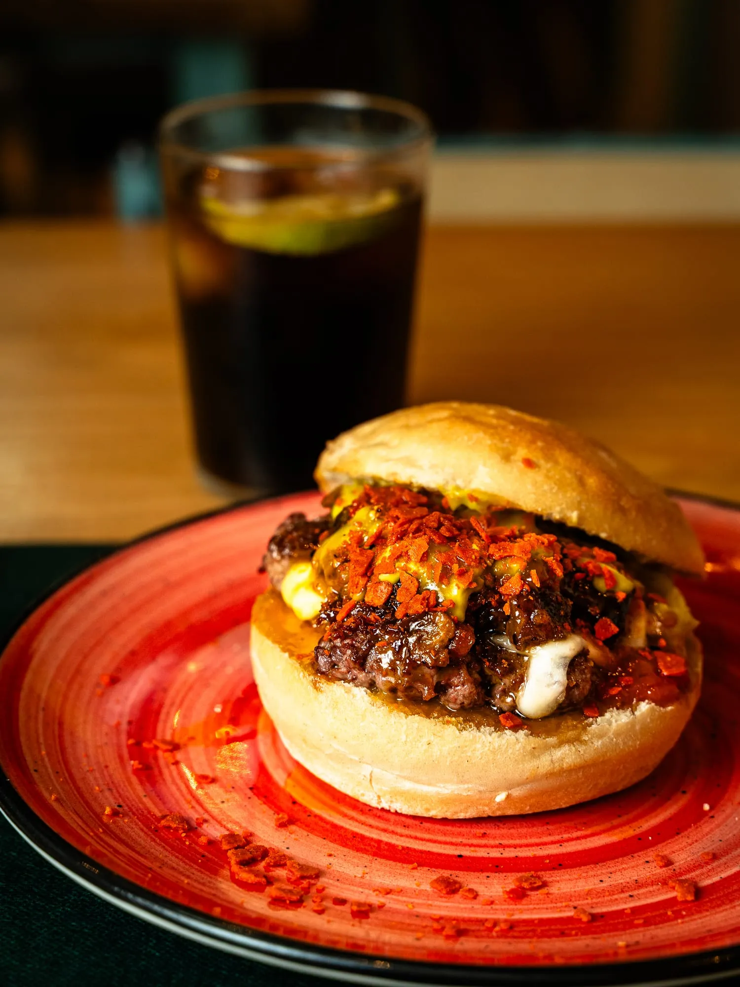 Hamburguesa en plato rojo con vaso de refresco al fondo, luz cálida