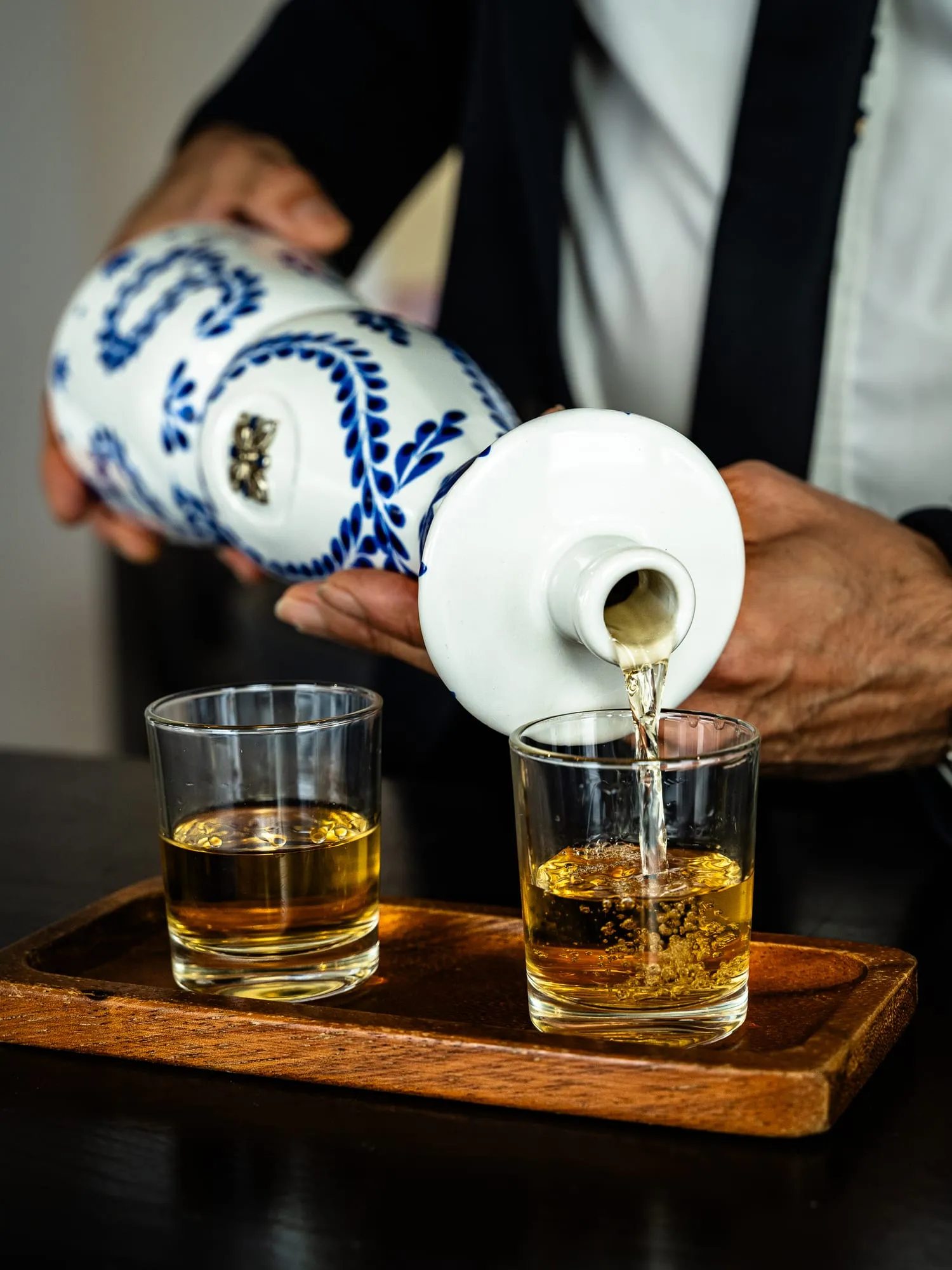 Manos sirviendo tequila desde su botella de cerámica en dos vasos bajos sobre bandeja de madera