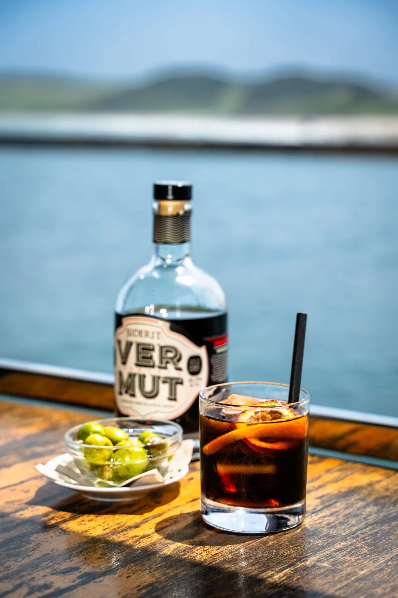 Botella de vermut Siderit con un vaso preparado y aceitunas frente al mar