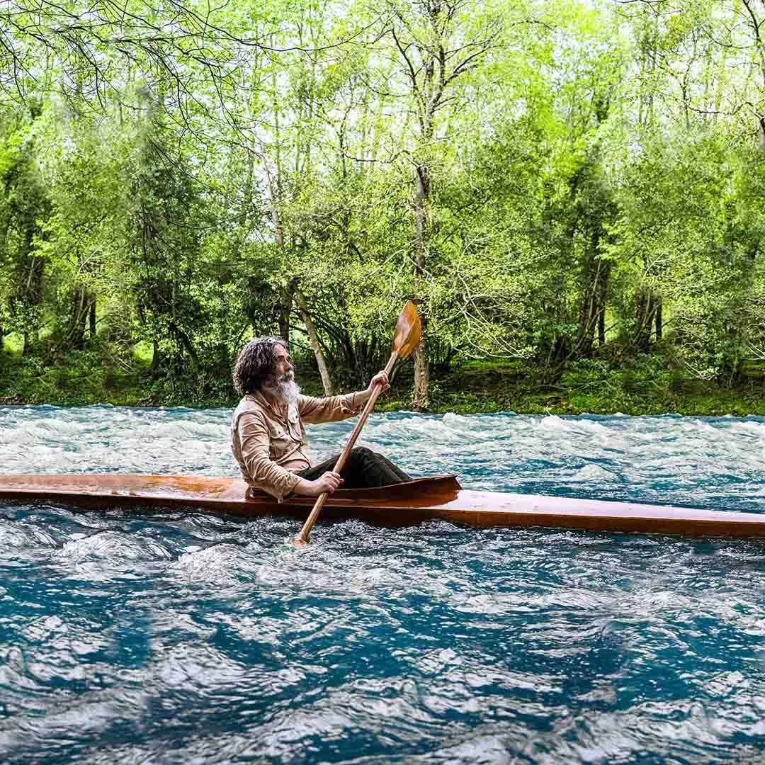 El protagonista rema en un kayak de madera por un río de aguas bravas entre árboles