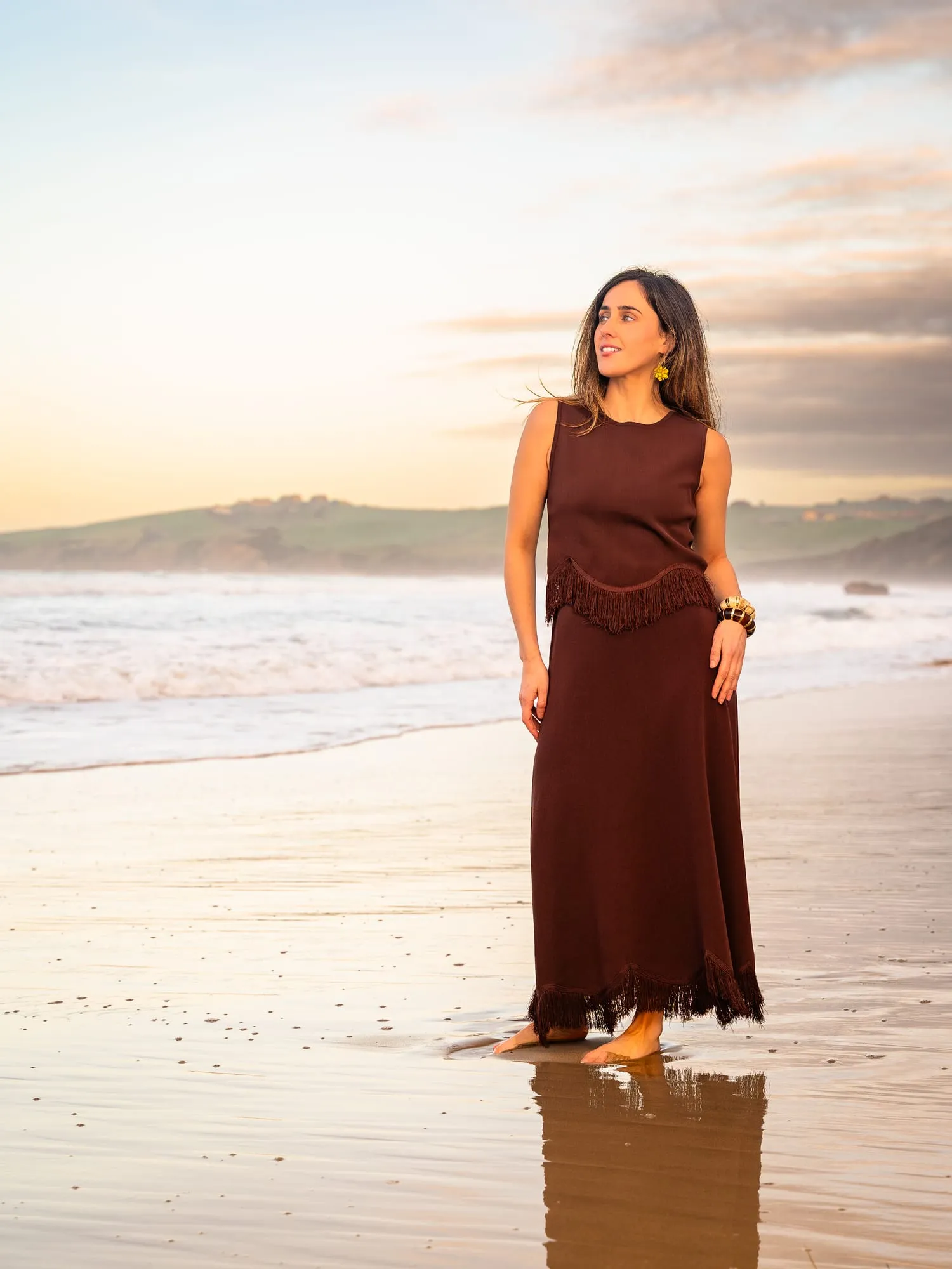 Modelo con vestido largo oscuro descalza en la orilla al atardecer, reflejada en la arena mojada