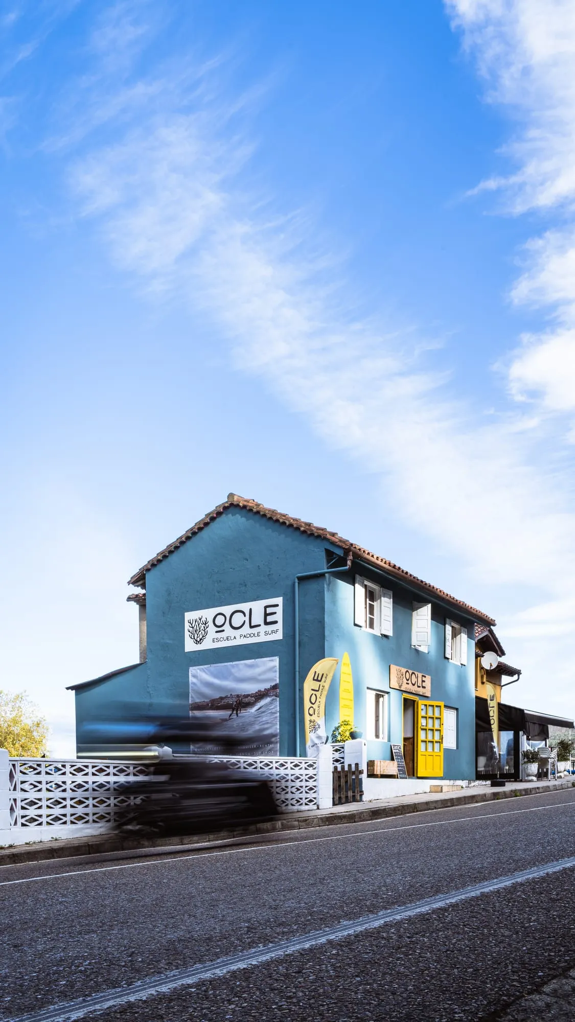 Fachada azul de la escuela Ocle Paddle Surf con el rótulo y la banderola amarilla