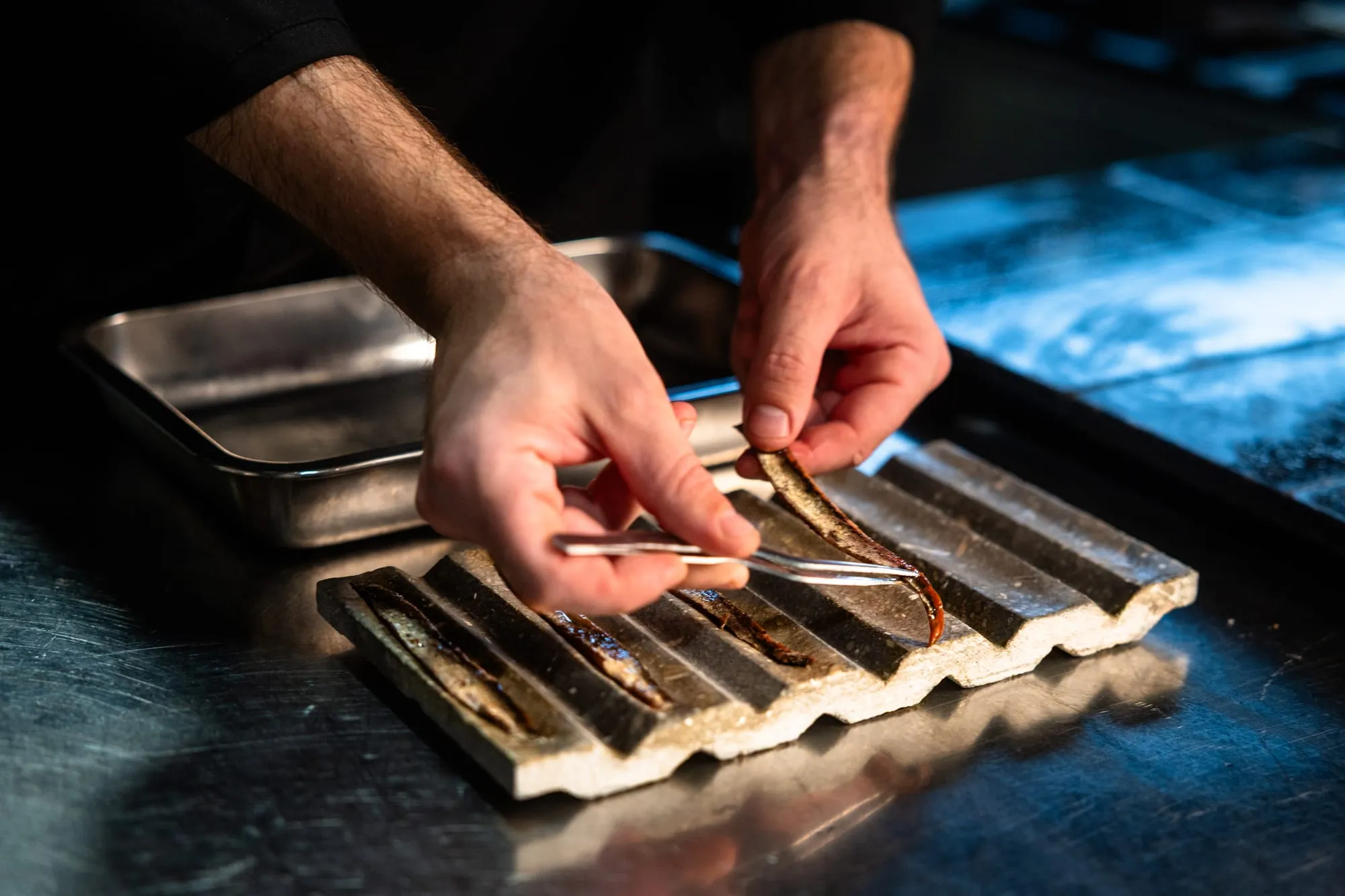 Manos de cocinero colocando filetes de anchoa con pinzas sobre una bandeja de acero