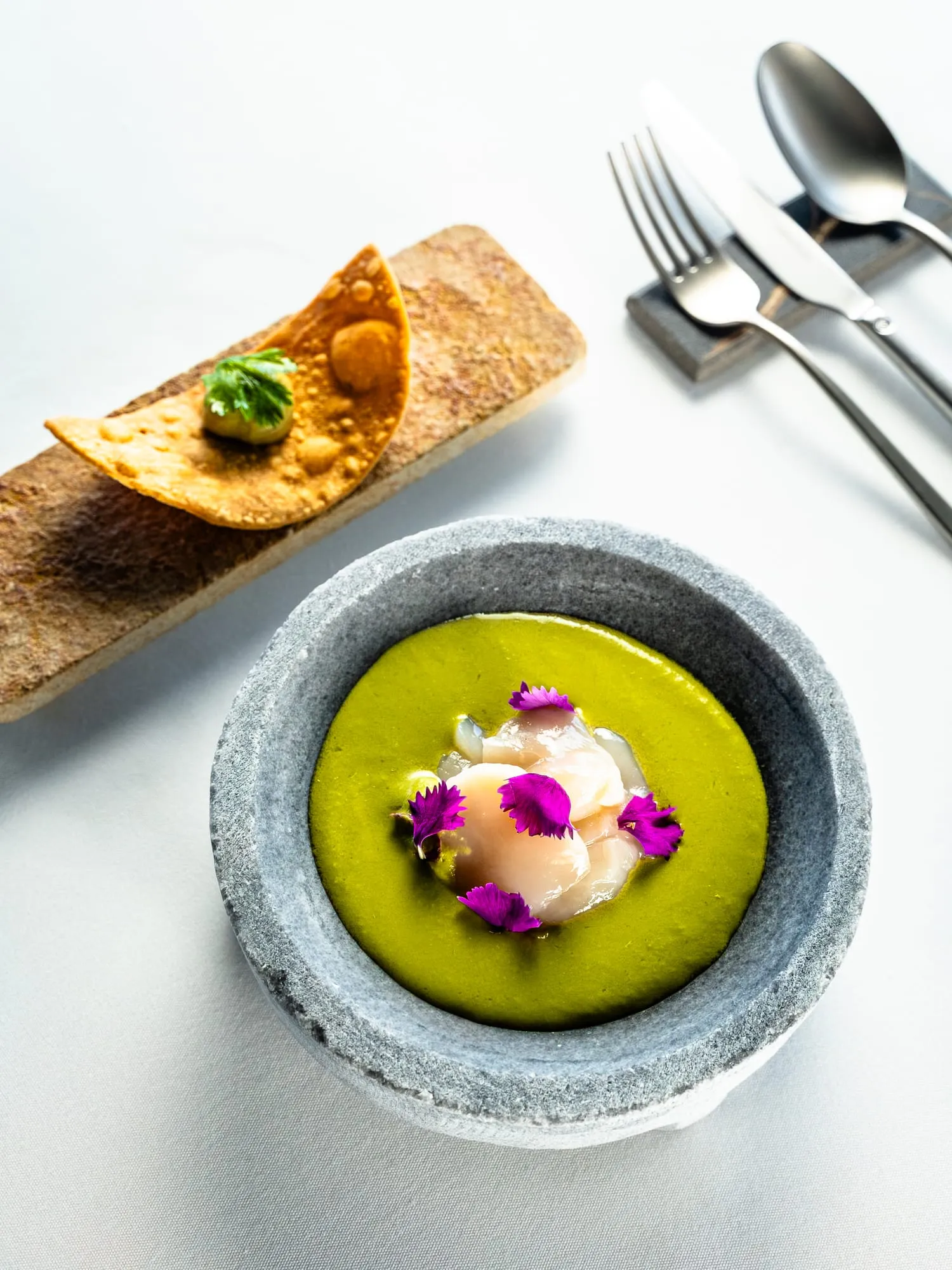 Crema verde con pescado y flores comestibles en cuenco de piedra, junto a una tostada