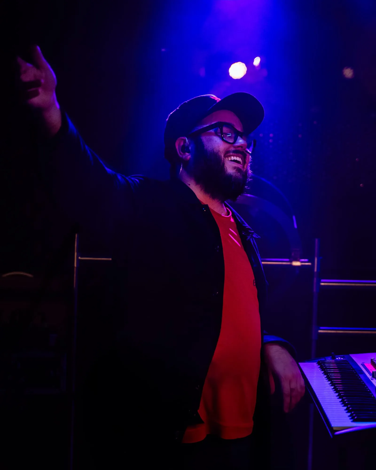 Él al teclado con gorra y camiseta roja, sonriendo y con el brazo en alto bajo luz violeta
