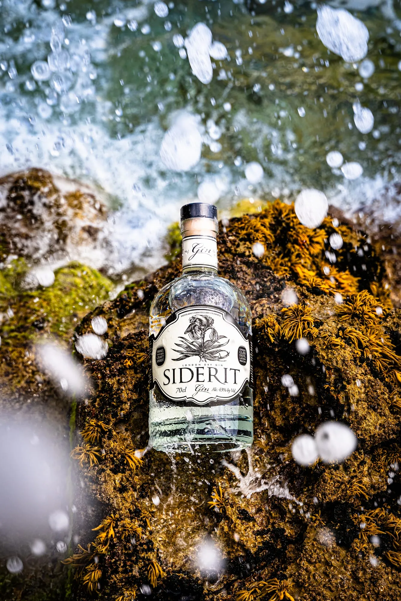 Botella de ginebra Siderit sobre rocas con algas mientras rompe una ola con salpicaduras congeladas