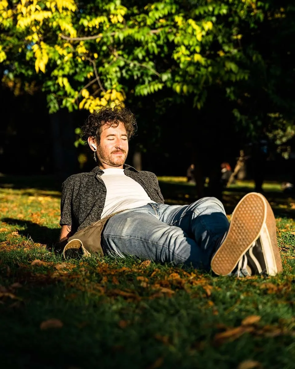 Hombre tumbado en la hierba de un parque con los ojos cerrados y auriculares, al sol de otoño